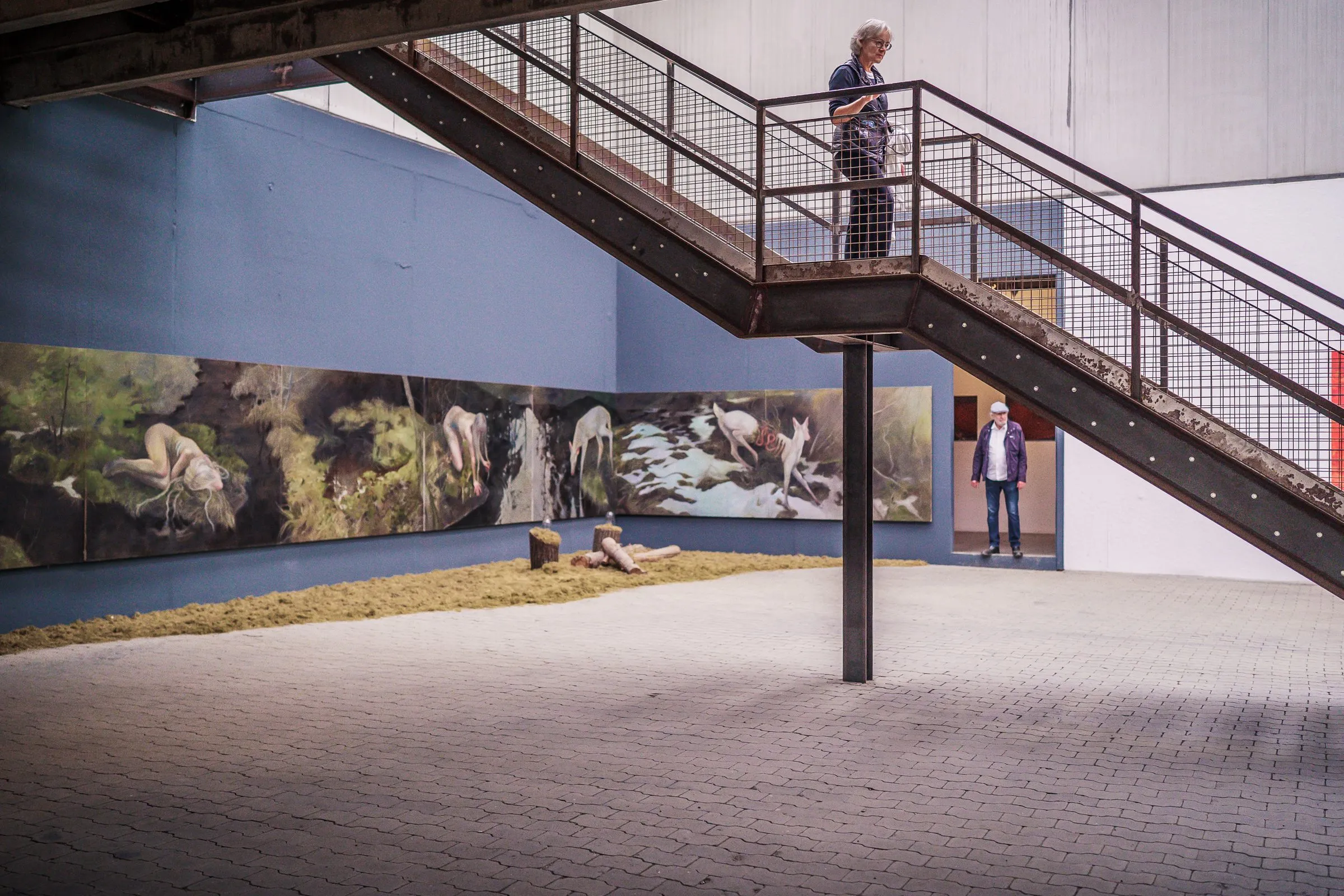 A long panoramic painting with forest and animals on blue walls, hay and a steel staircase in front