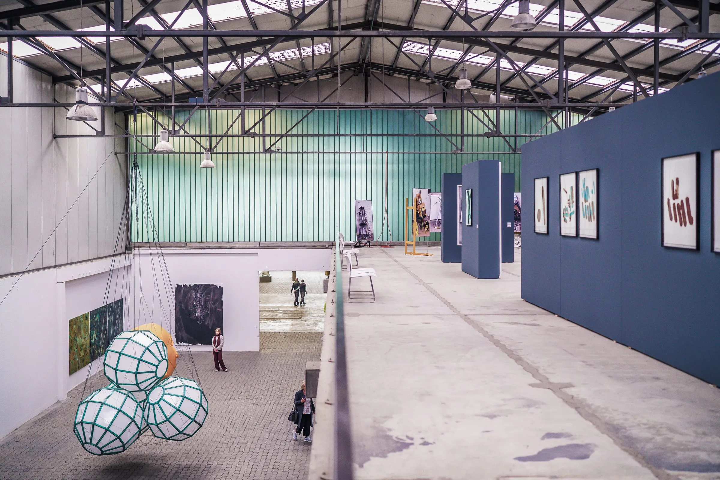 A view from a gallery into the hall, hanging lattice spheres on the left, framed pictures on blue walls on the right