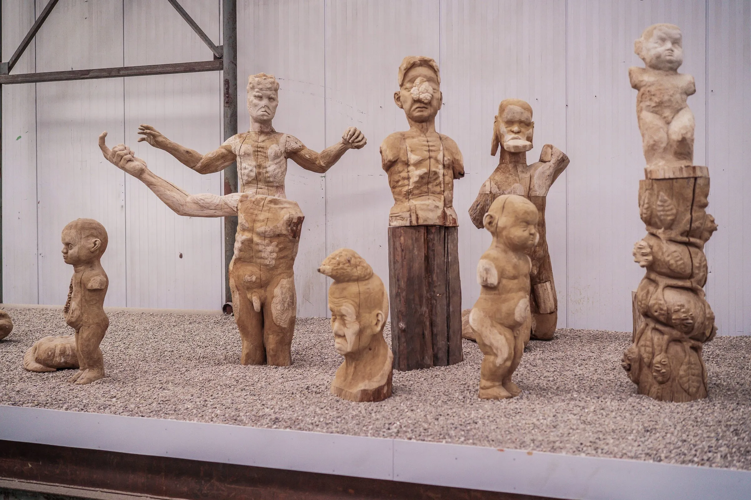 A group of carved wooden figures, people and animals, on a gravel bed in front of a pale wall
