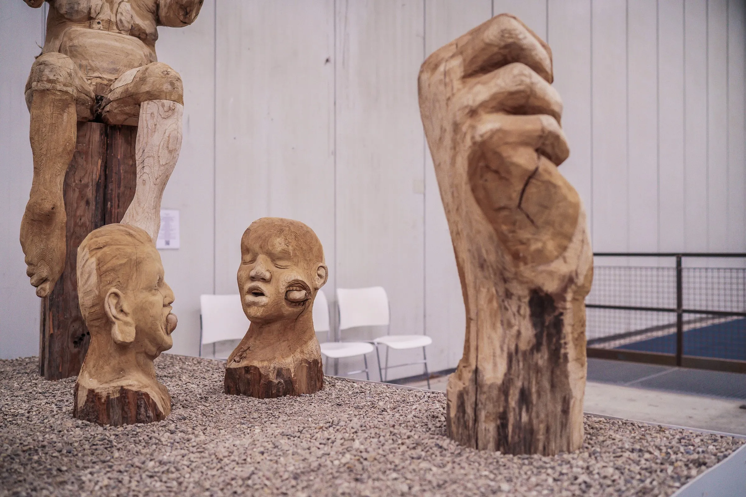 Carved wooden heads and a large wooden foot on gravel, white chairs behind