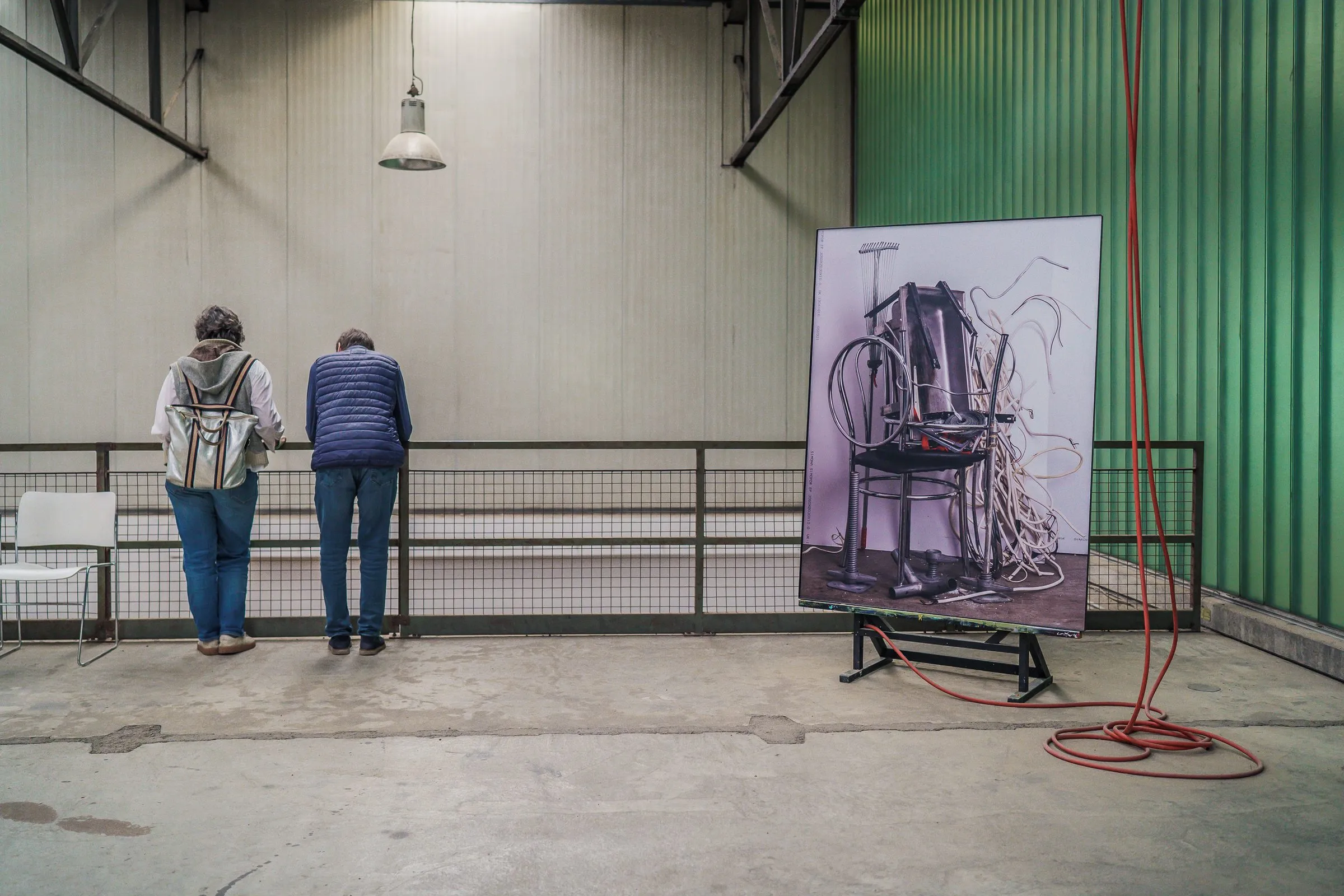 Two visitors lean on the railing, beside them a picture with a chair and cables, a red cable on the floor in front