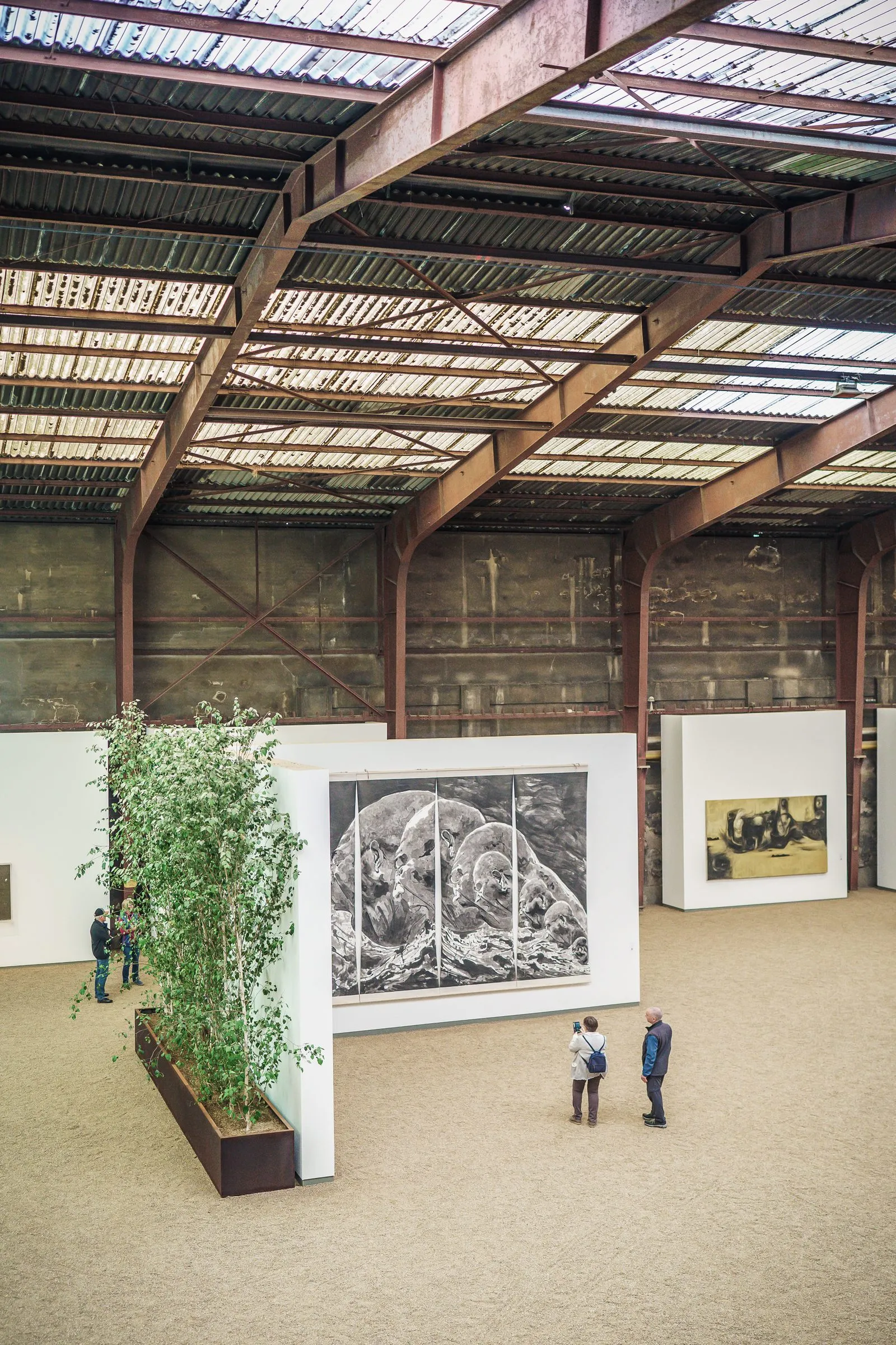 Portrait: a view from above onto a hall with bamboo in planters, large drawings and two visitors