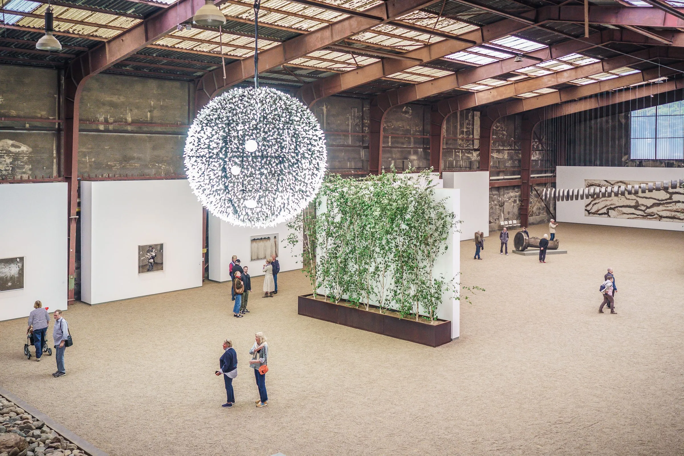 A huge industrial hall from above, a glowing sphere of light in the middle, bamboo in planters, white walls and visitors spread out