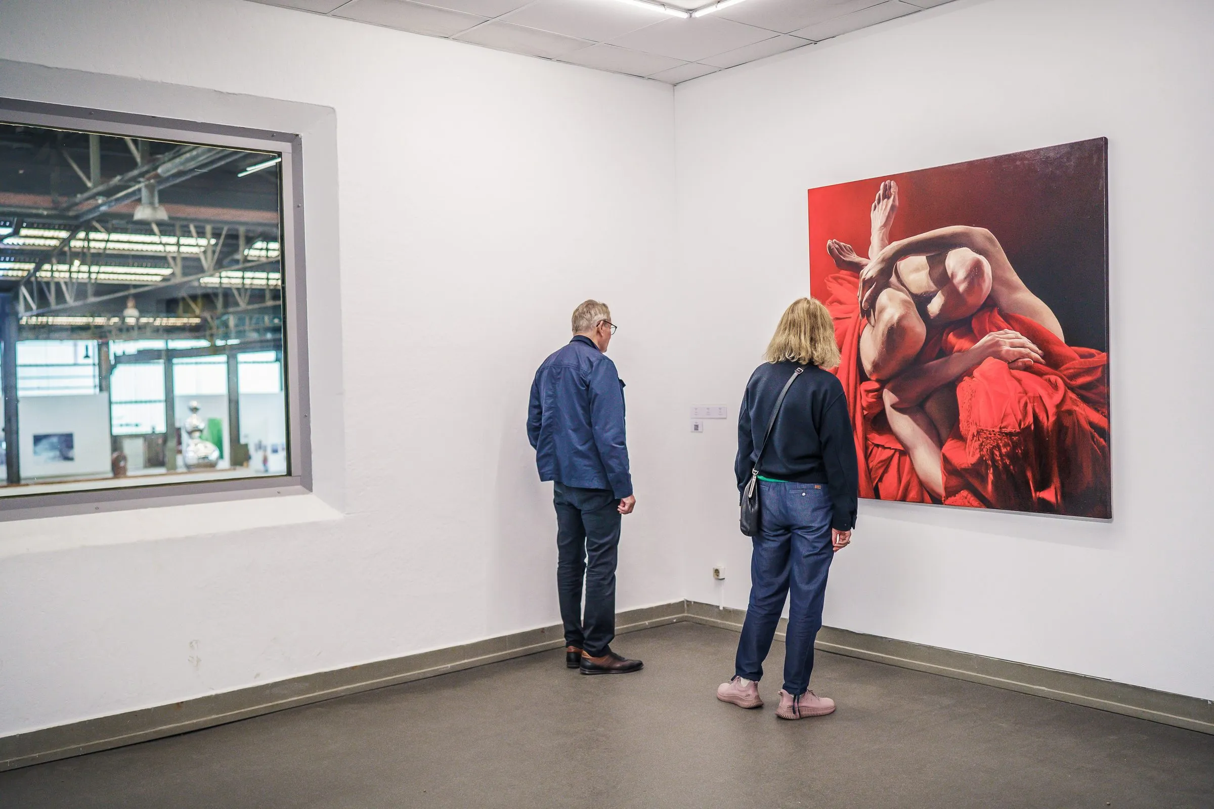 A couple stands in front of a painting with bodies in red fabric, a window into the hall on the left