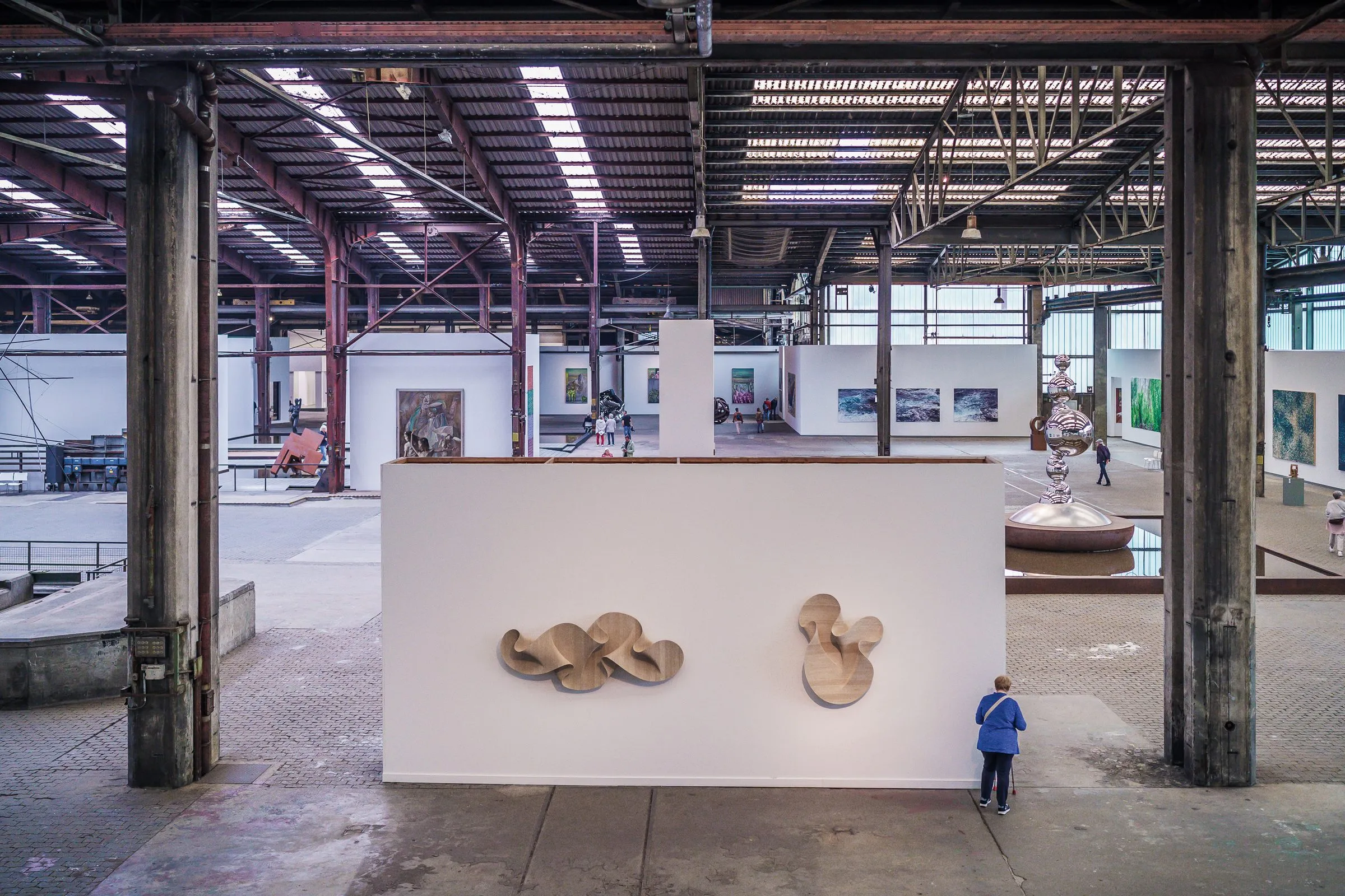A white wall with two curved wooden sculptures between old steel columns, a visitor next to it