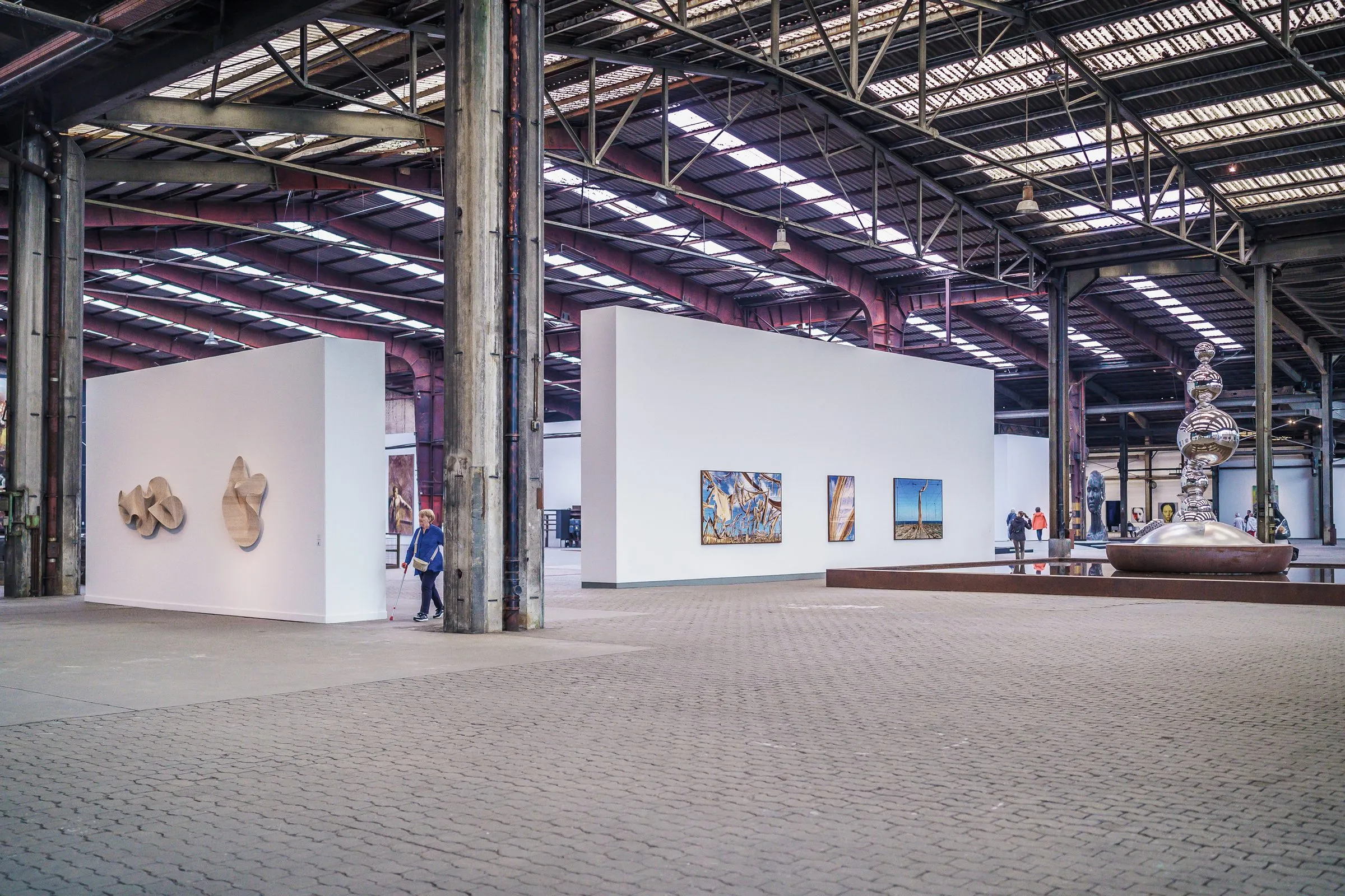 A wide hall with steel columns and skylights, white exhibition walls with sculptures and pictures