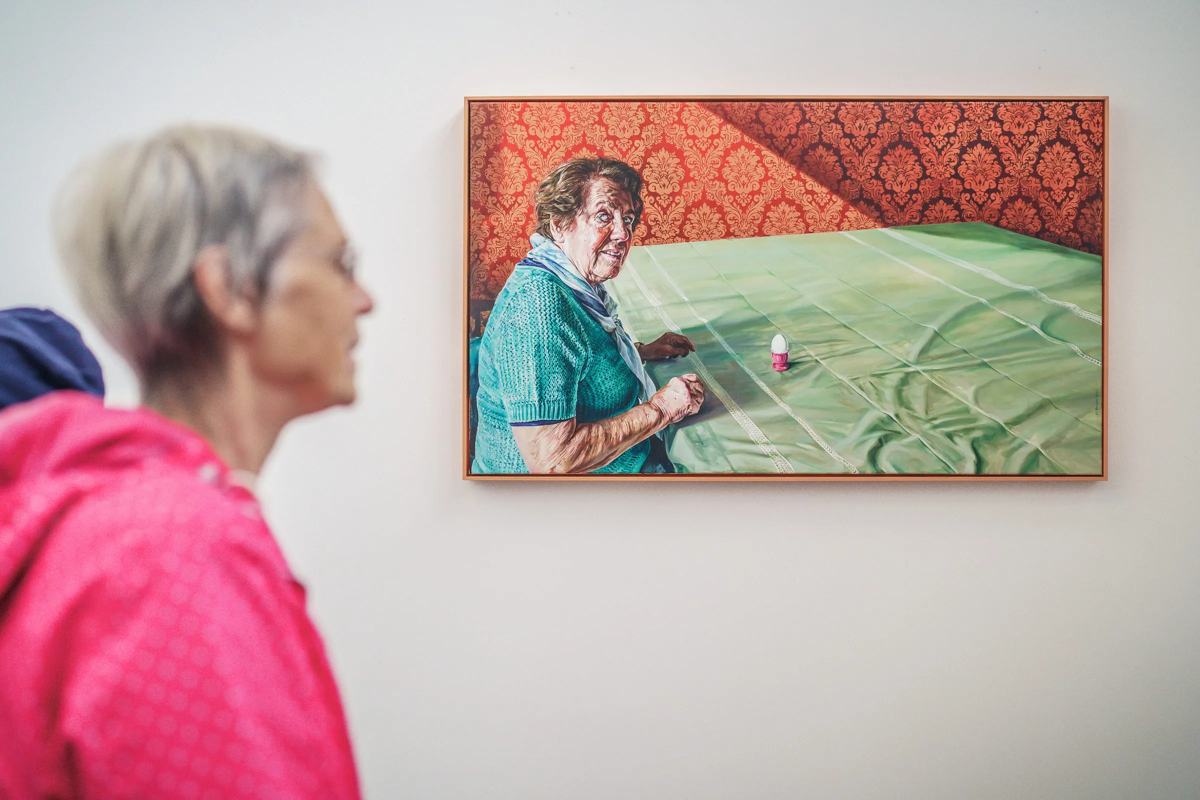A visitor in profile in front of a painting of an older woman at a green table against red wallpaper