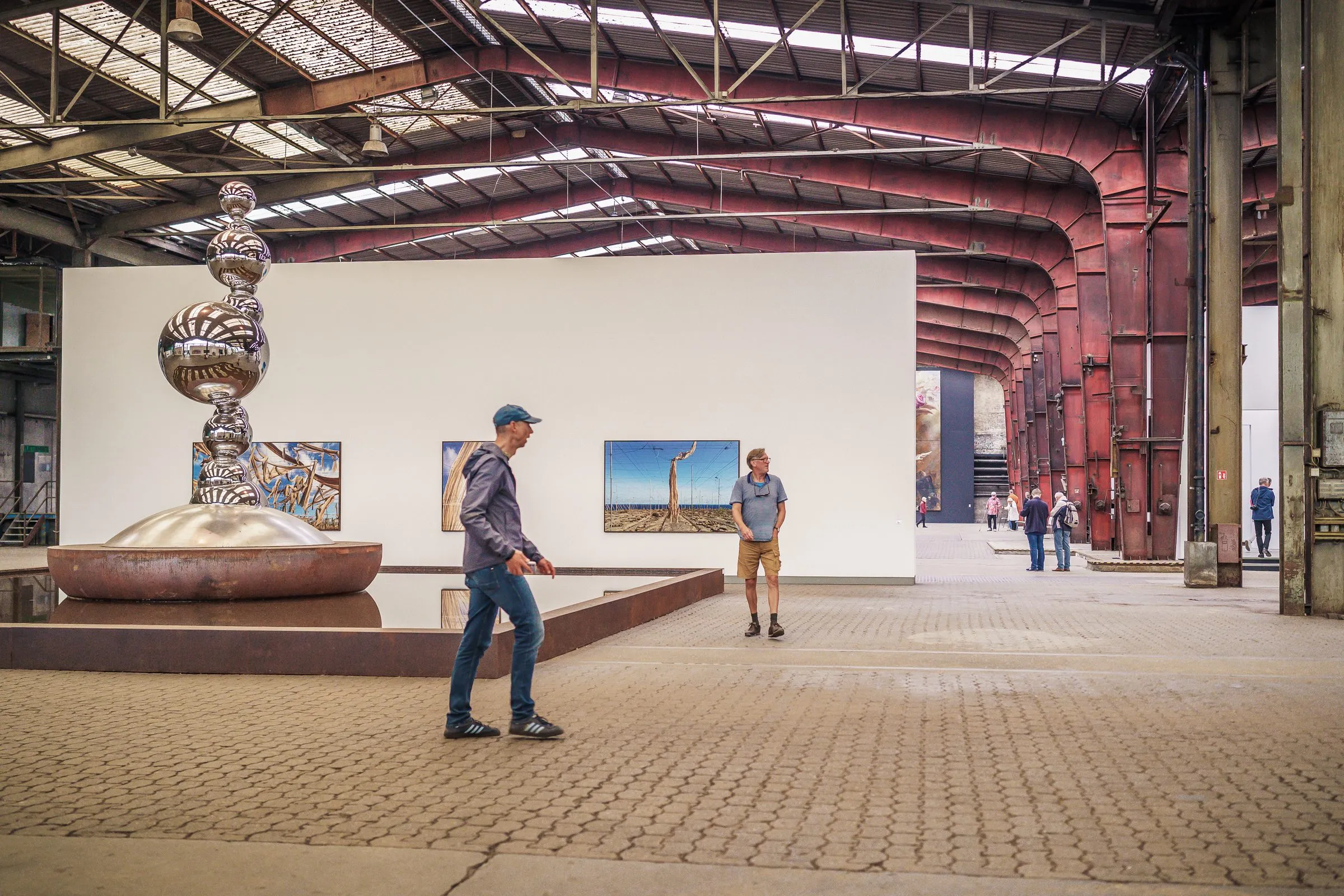 A silver stacked sculpture in a shallow water basin, two visitors in front, pictures on a white wall behind