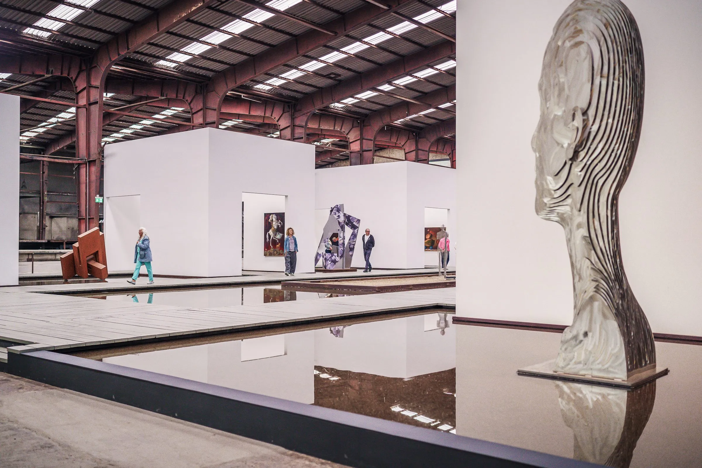 A large head in profile made of silver metal layers at the edge of a water basin, the hall behind