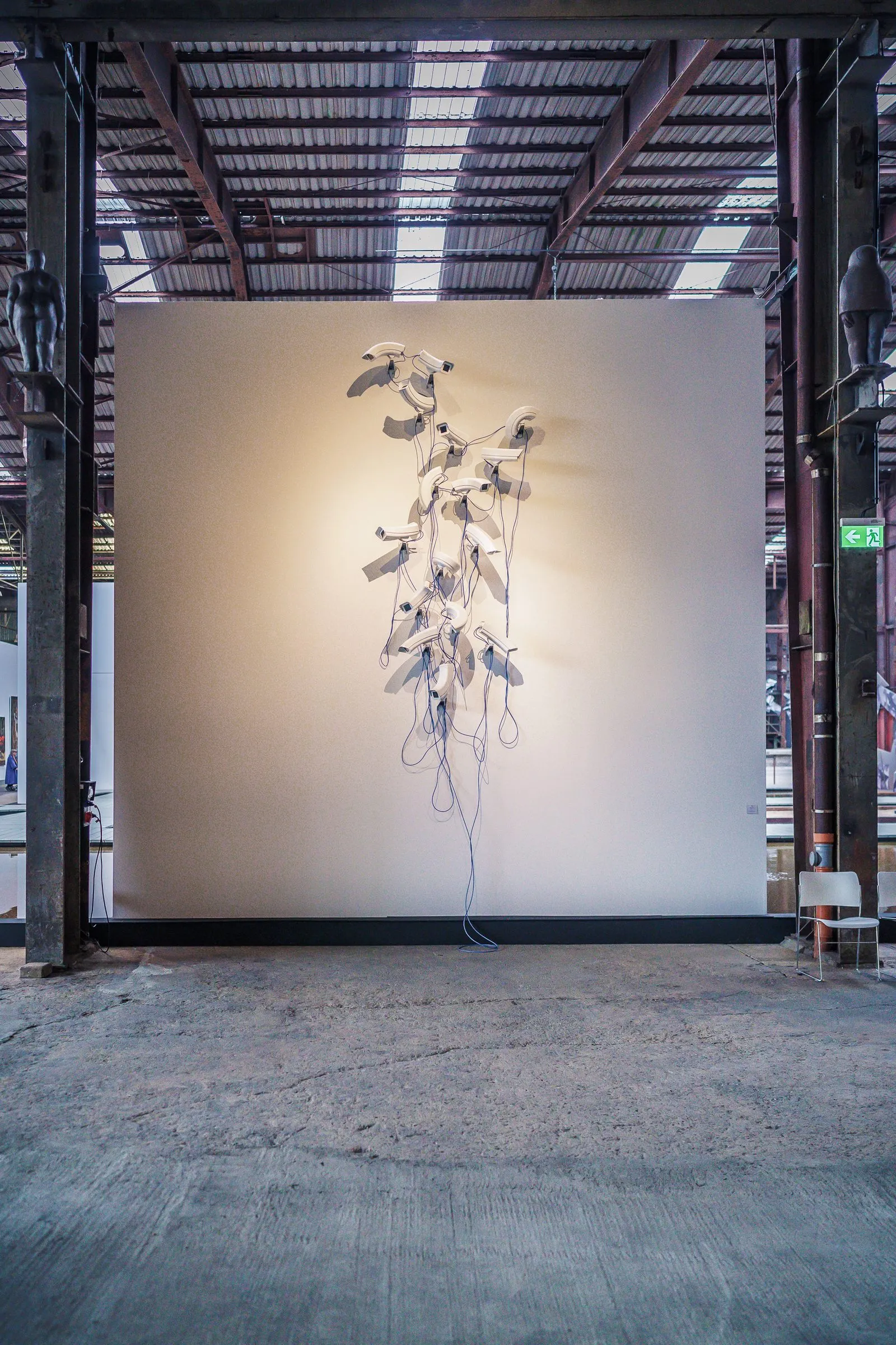 Portrait: a pale, tendril-like sculpture hangs in front of a lit white wall between steel columns