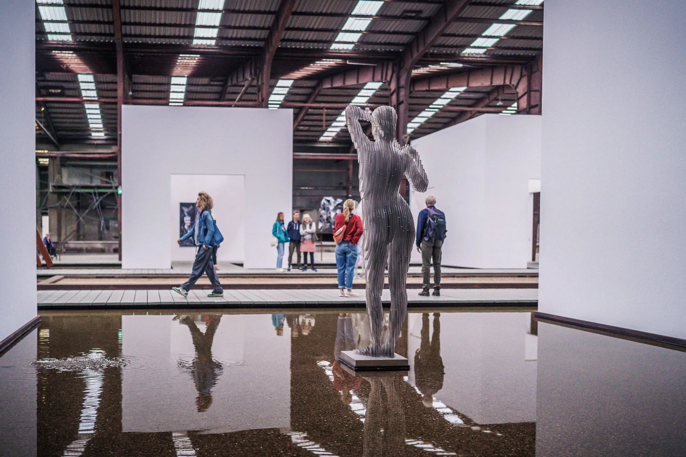 A standing figure on a walkway in the water basin, its reflection below, visitors in the background