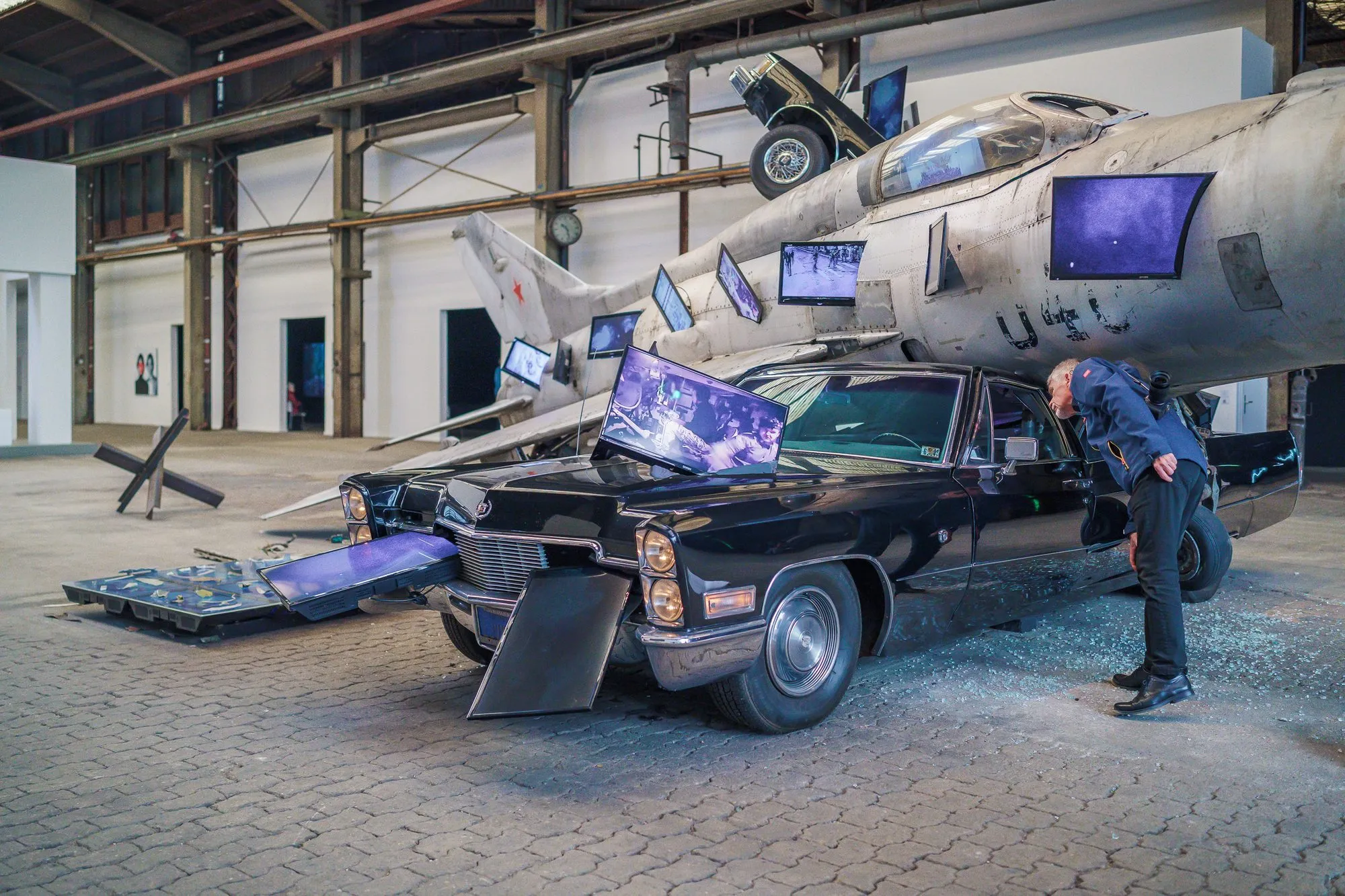 A large installation of a fighter jet, a black car and screens, a visitor leaning over it