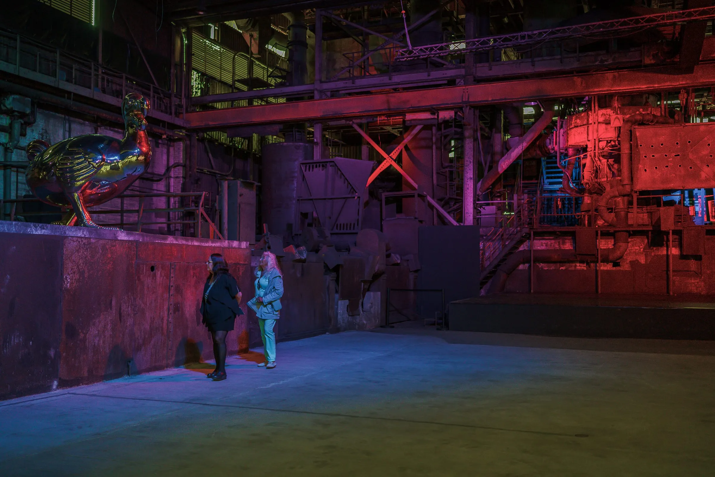 A dark hall in red light with old industrial structures, an animal sculpture above, two visitors below