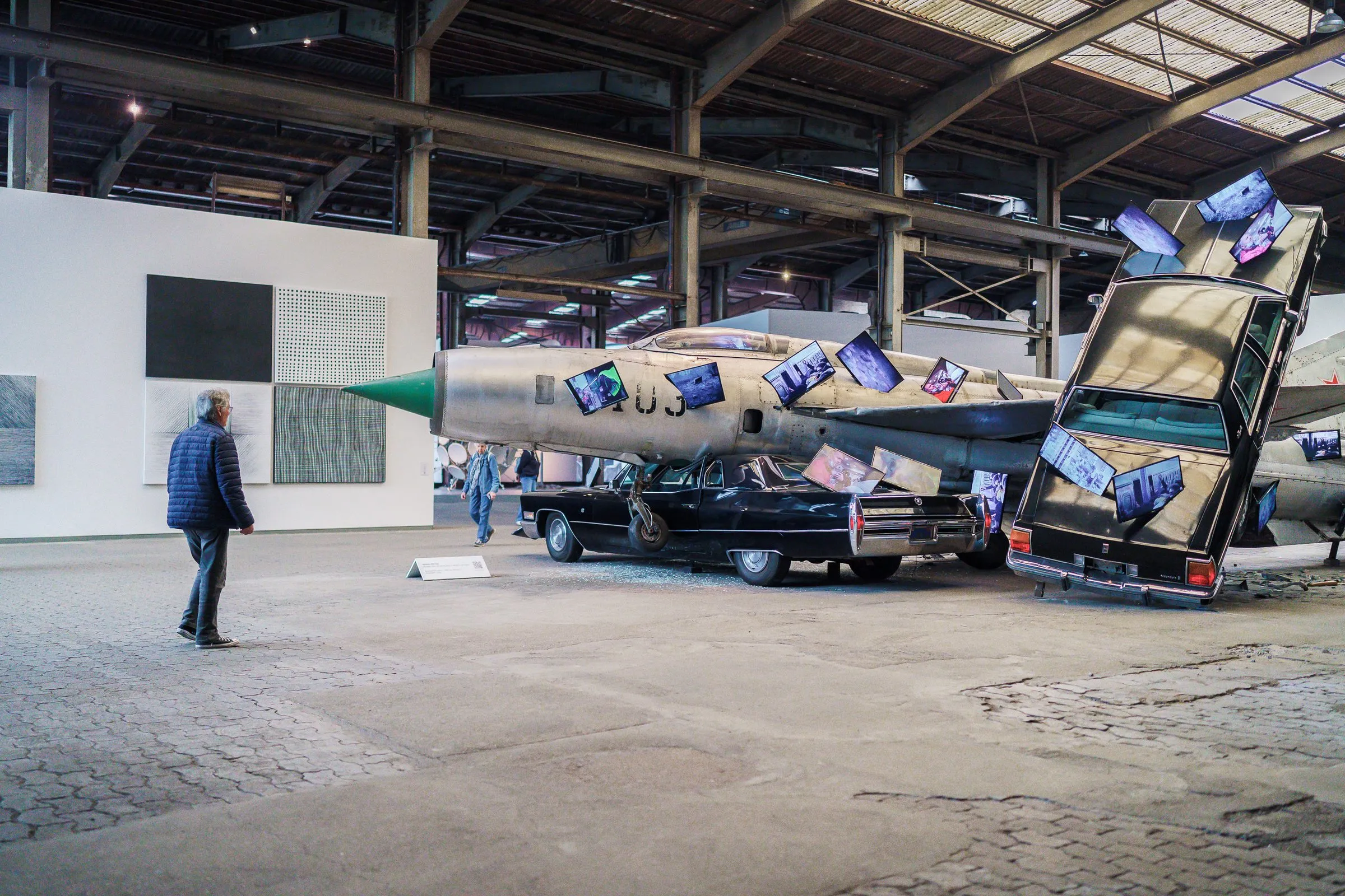The installation with fighter jet and car from a distance, a visitor in front, pictures on the wall behind