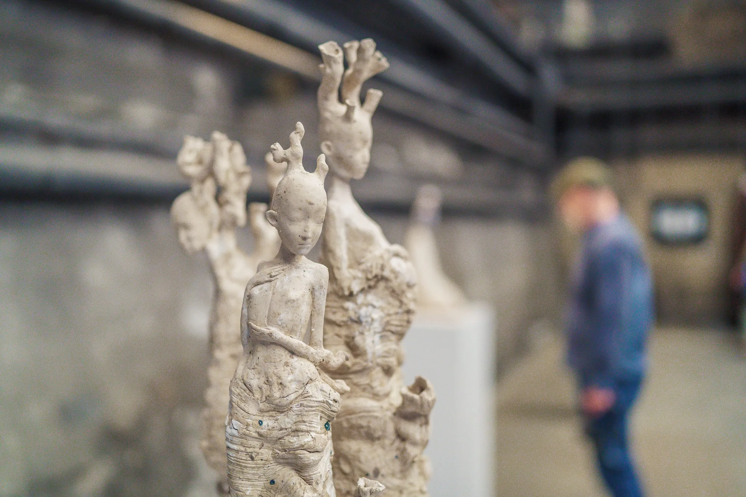 Close-up of a pale, branching ceramic sculpture with figures, a visitor out of focus in the background