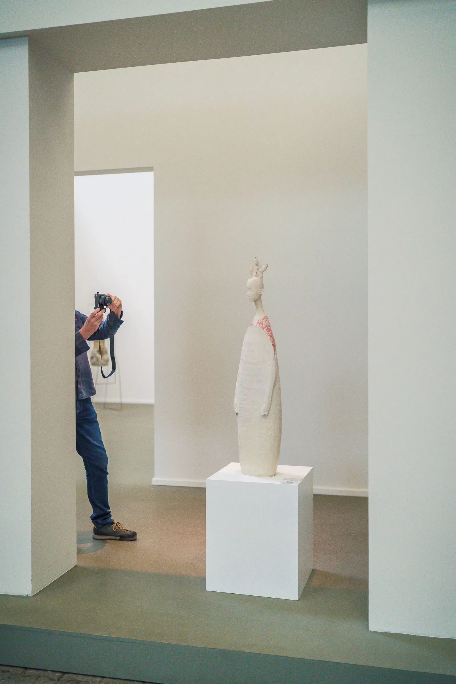 Portrait: a pale figure on a white plinth in a white room, someone photographing beside it