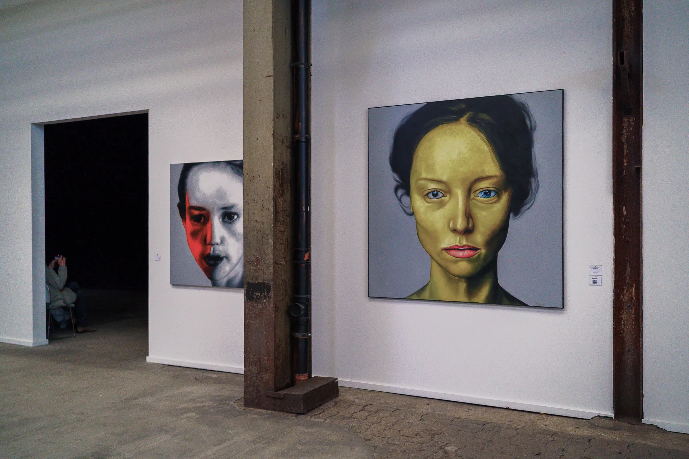 Two portrait paintings on a white wall, a woman with a yellowish painted face and a half-covered face