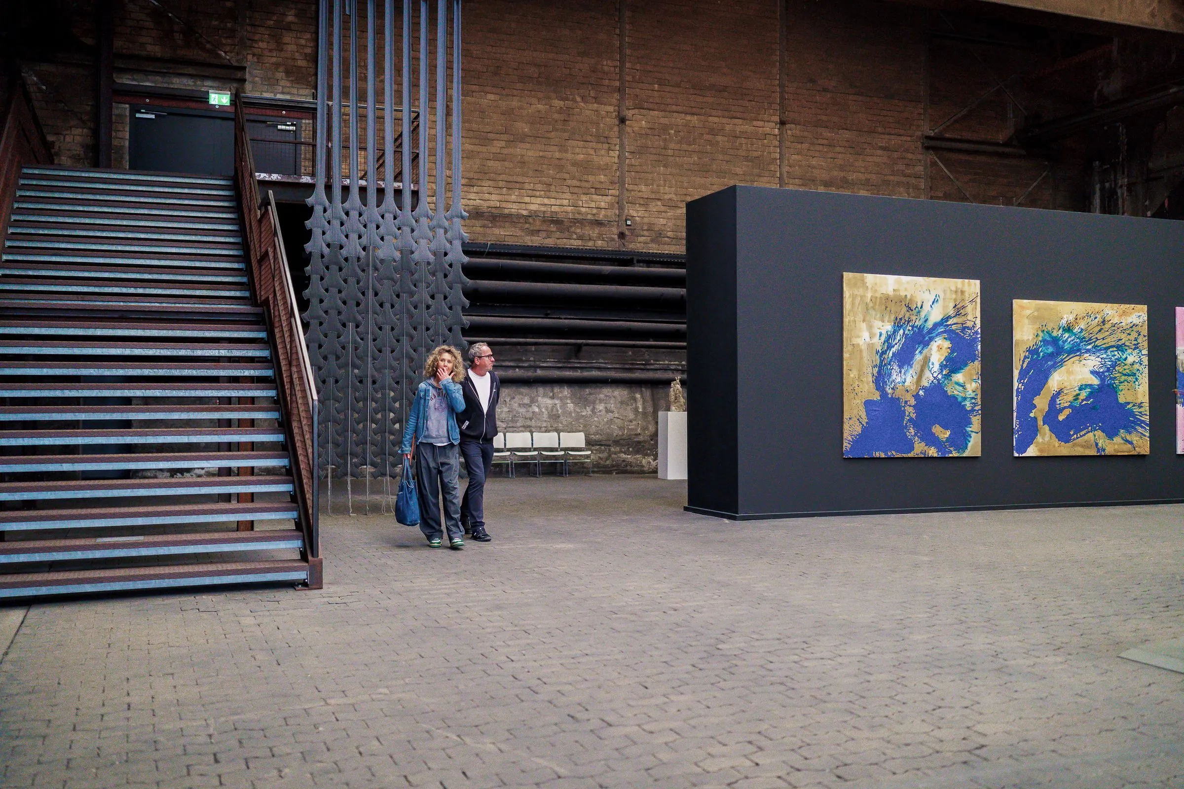 A steel staircase and a black wall with pictures in blue and gold, two visitors in front