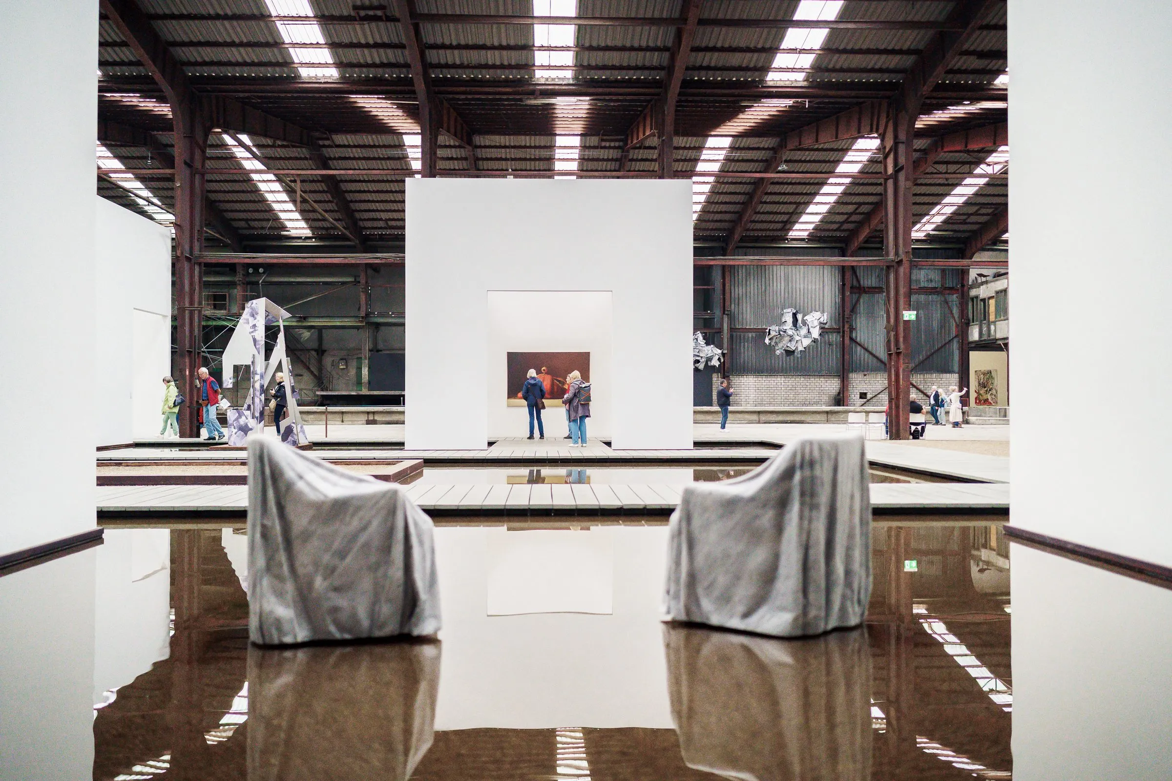 Two white-draped armchairs stand in the water, a white room with a picture and visitors behind