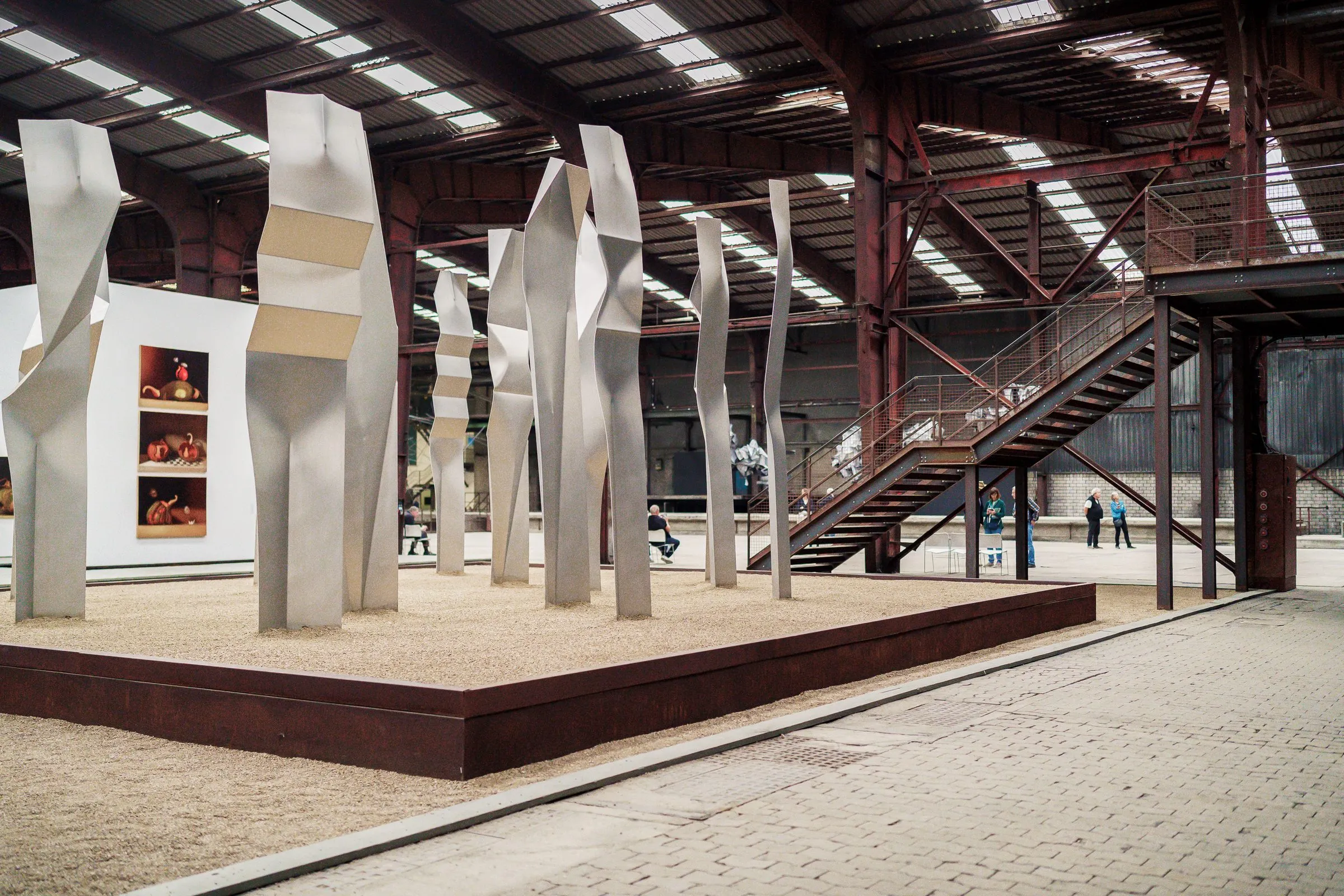 Tall white folded sculptures on a platform, a steel staircase in the hall beside them