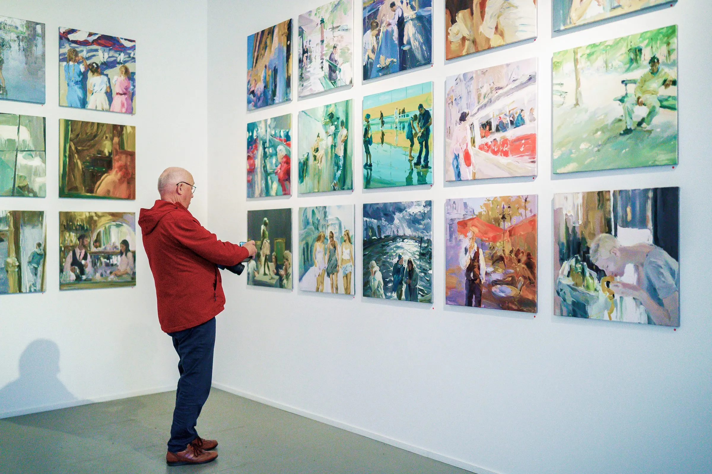 A man photographs a wall full of smaller pictures in light colours