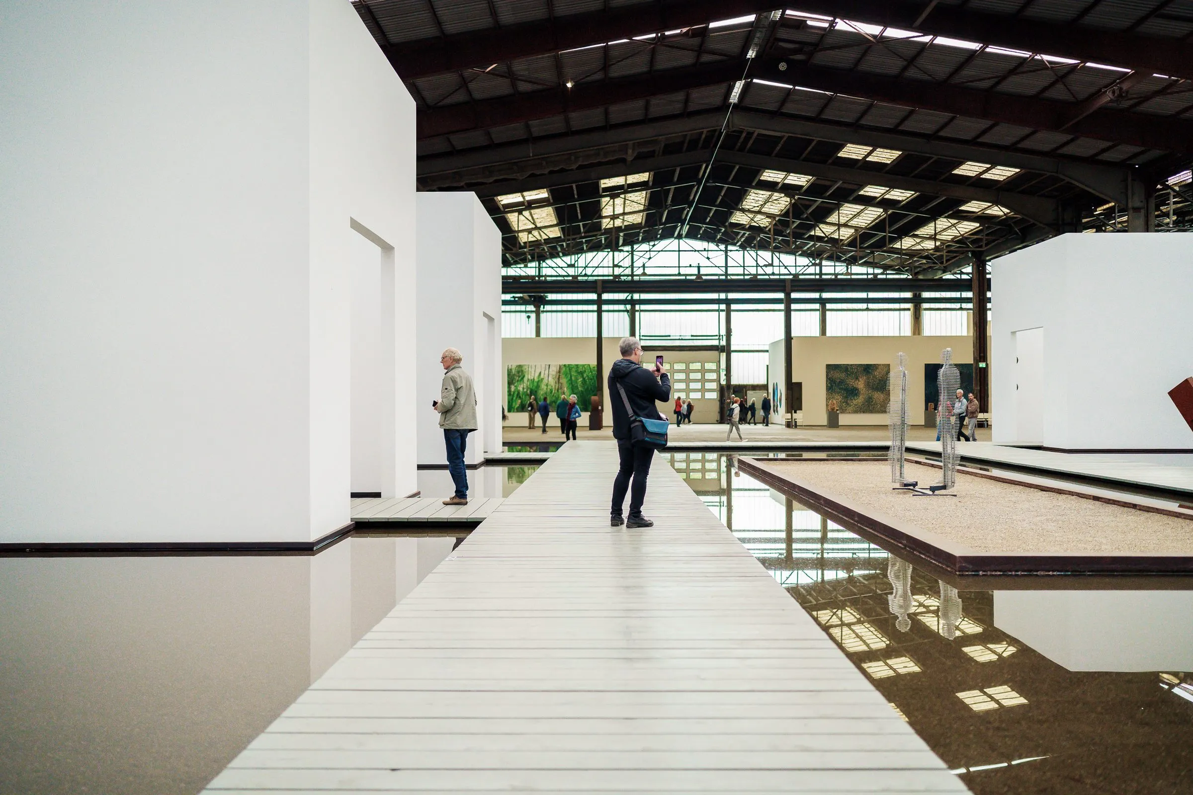 A pale wooden walkway leads over a water basin between white walls, two visitors at the end