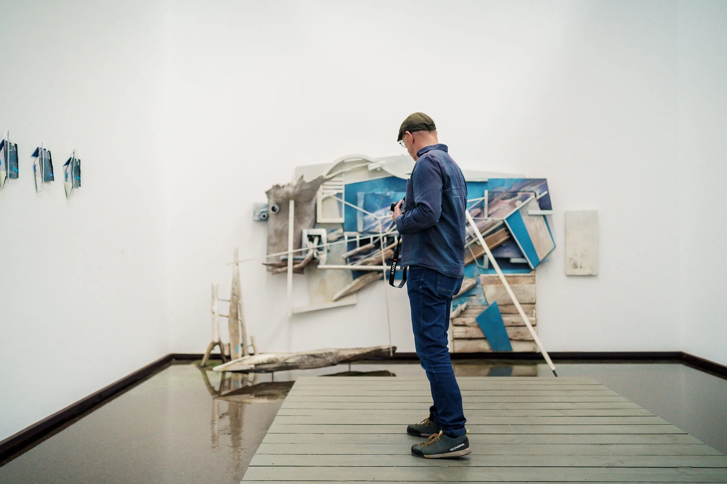 A man stands on a walkway in front of a wall installation of boards and picture fragments