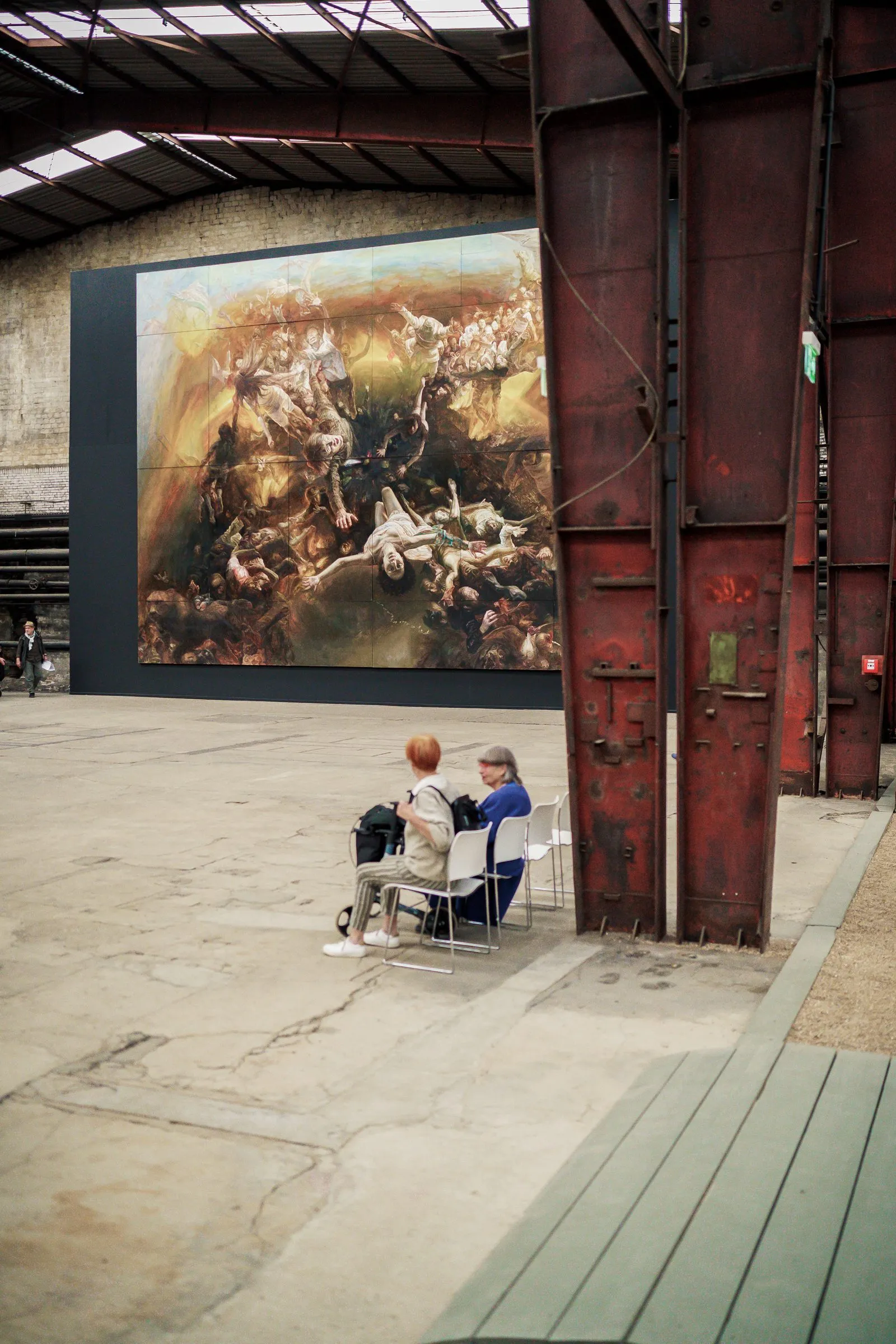 Portrait: two visitors sit small in front of the huge painting, a rusty steel girder beside them