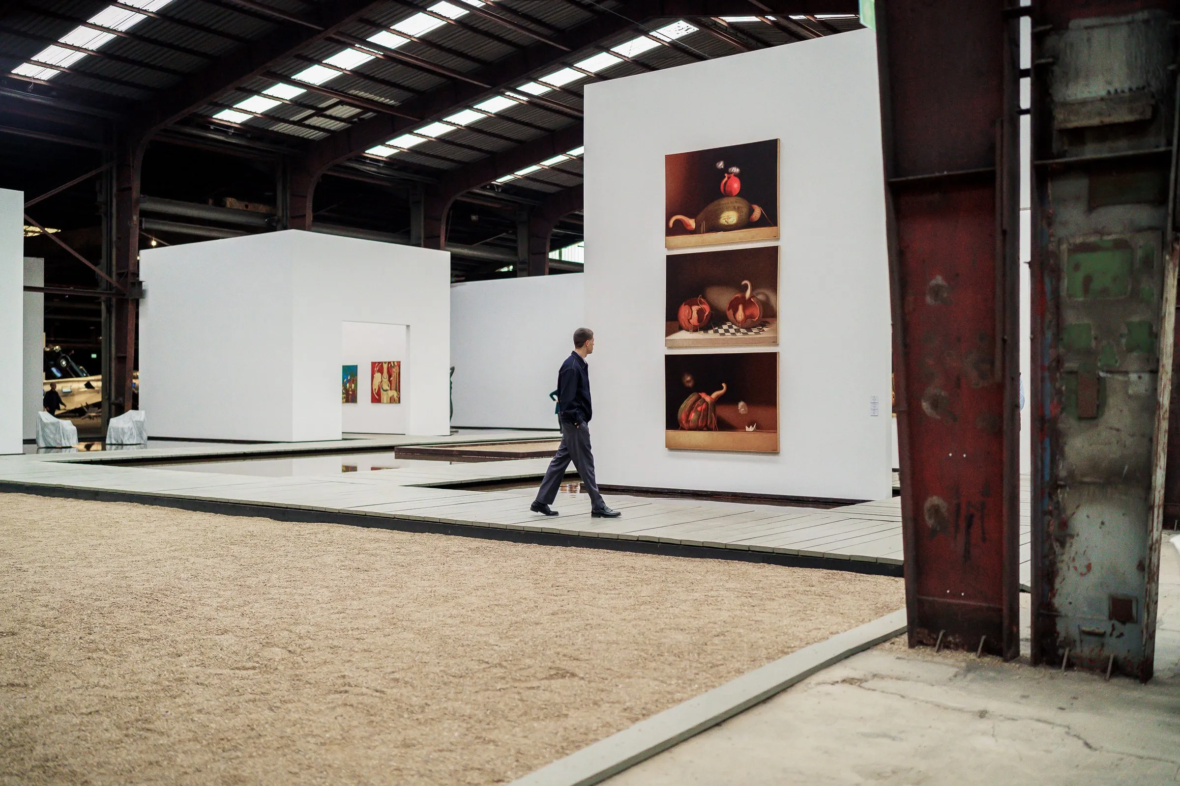 White exhibition cubes on gravel, a man walking past, a picture with red fruit in an opening