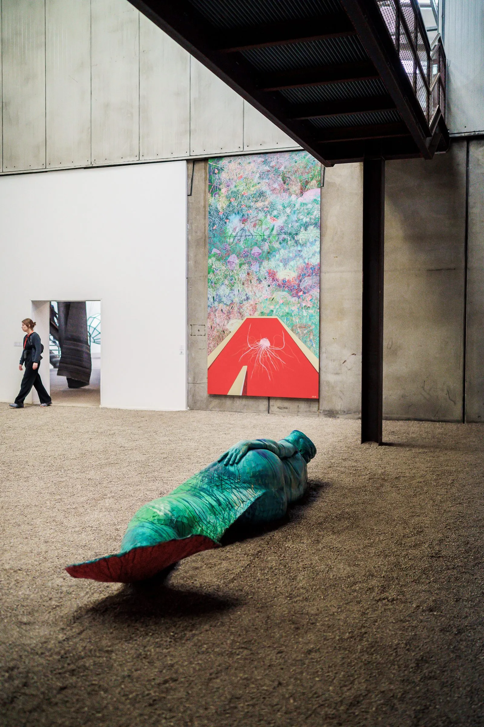 Portrait: a reclining, green-wrapped figure on a red base on the hall's sand floor, a picture behind