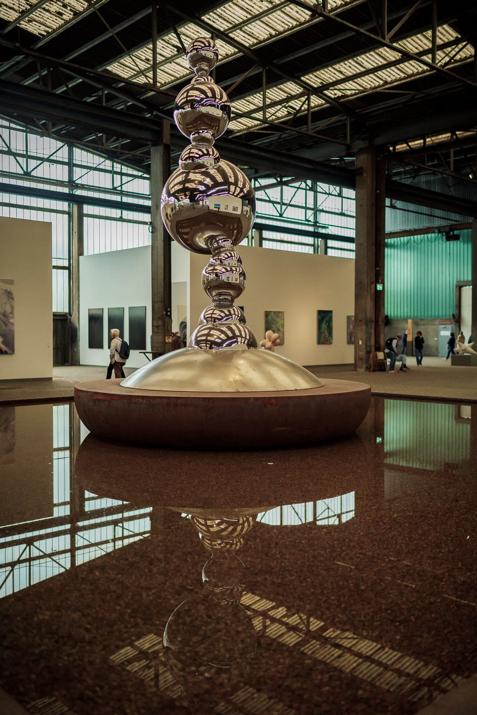 Portrait: a tall silver stacked sculpture reflected in the water basin