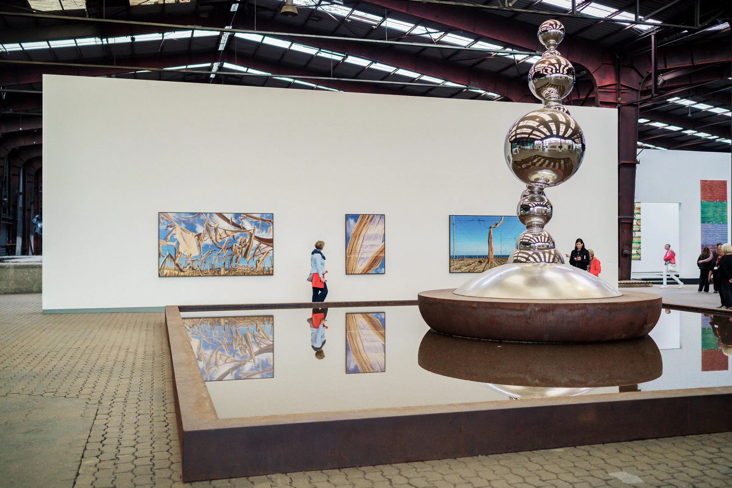 The silver stacked sculpture in the water basin in front of a white wall with pictures, visitors in front