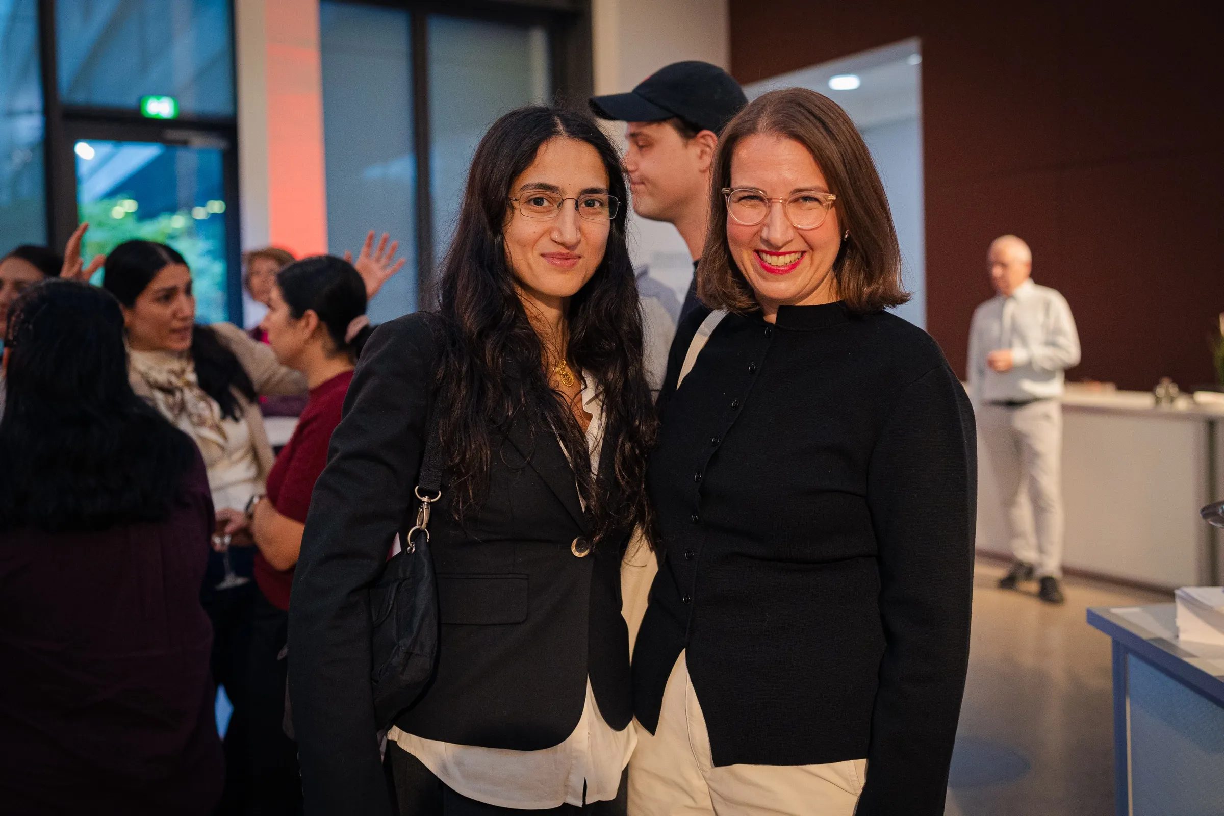 Leyla Yenirce and Dr. Anna Heinze in the foyer smiling into the camera, guests and the red light of the evening behind them