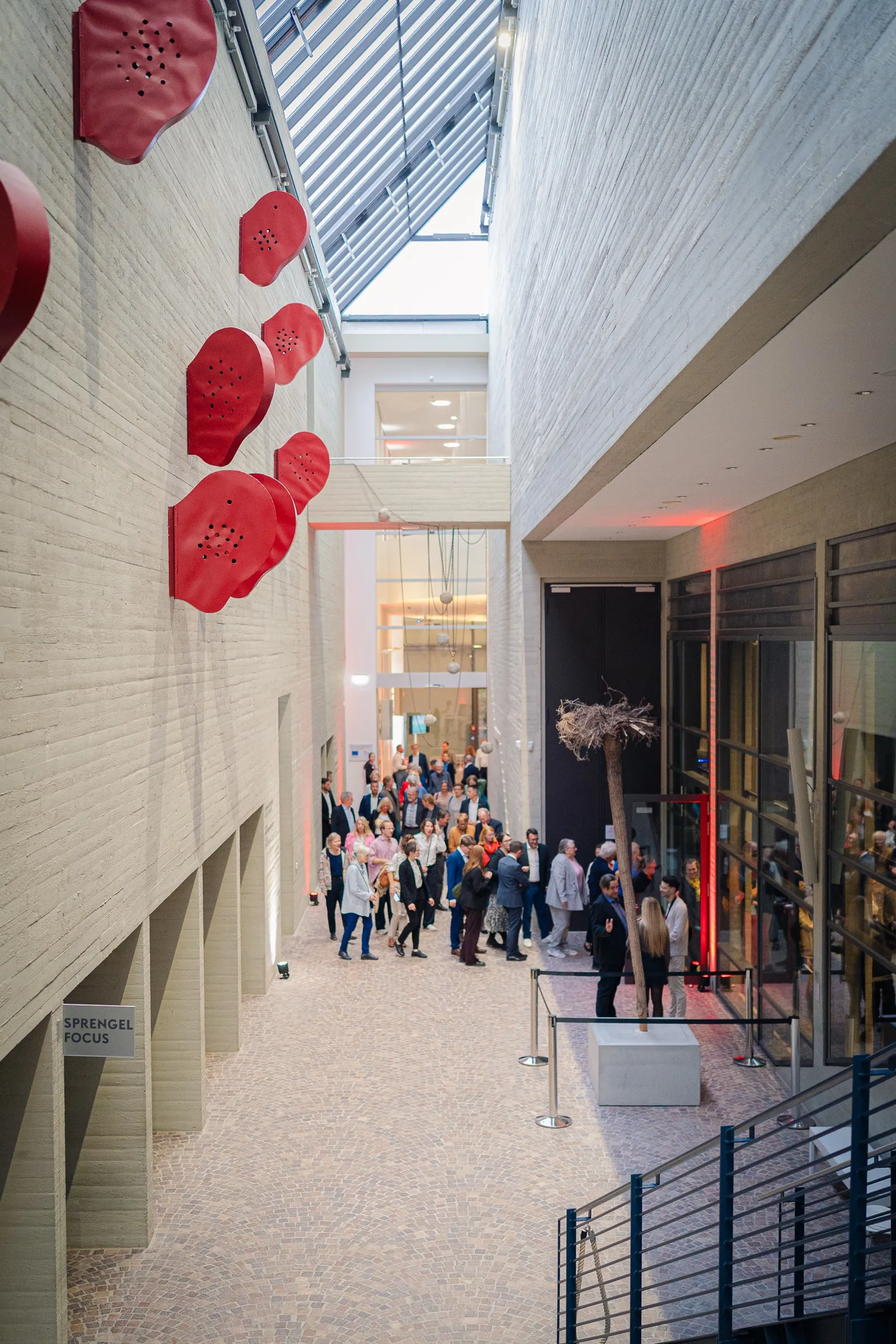 Portrait format: a narrow, high corridor with red objects on the pale wall, guests streaming in below