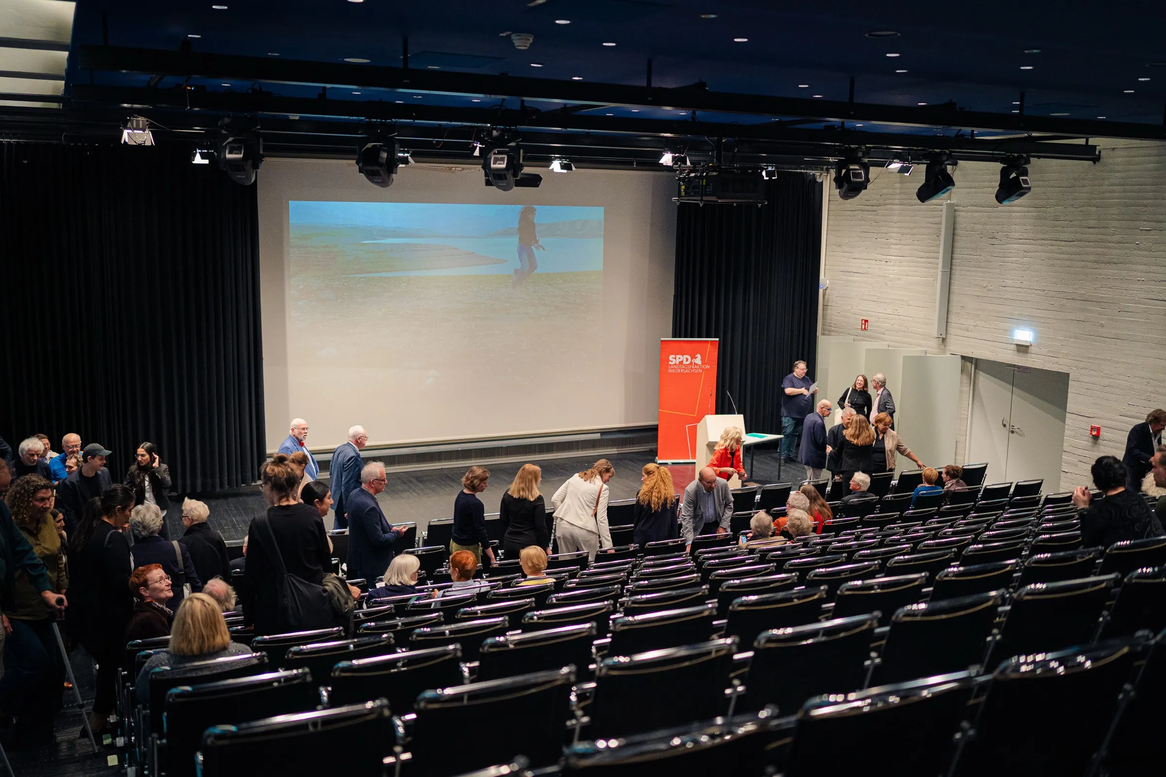 The hall from the back, black rows of chairs, the screen at the front and a red banner of the SPD parliamentary group