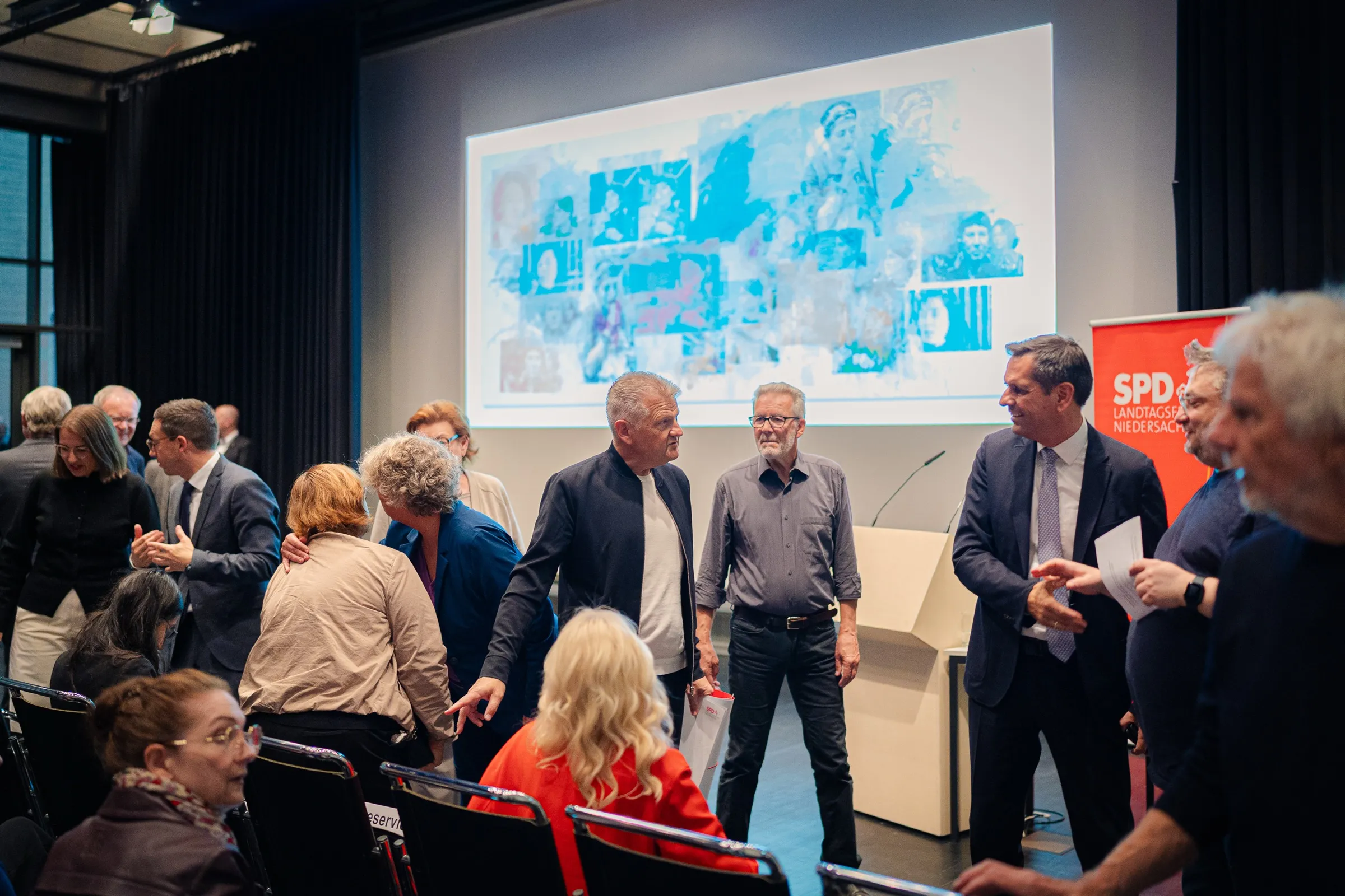Guests talking in front of the stage, on the screen a large painting with blue areas and many faces