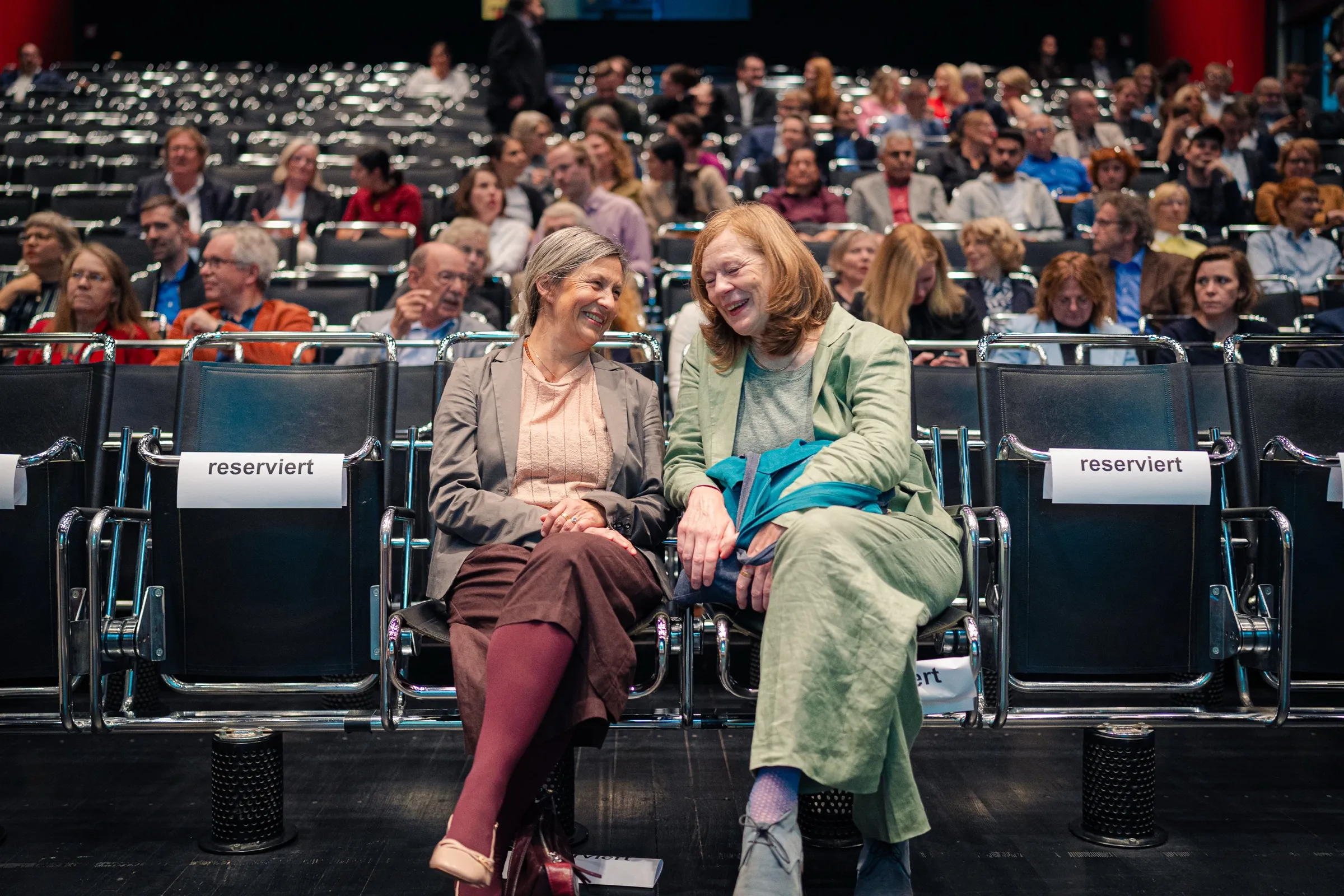 Anne Prenzler and another woman laughing on chairs marked “reserviert”, the full stands behind them