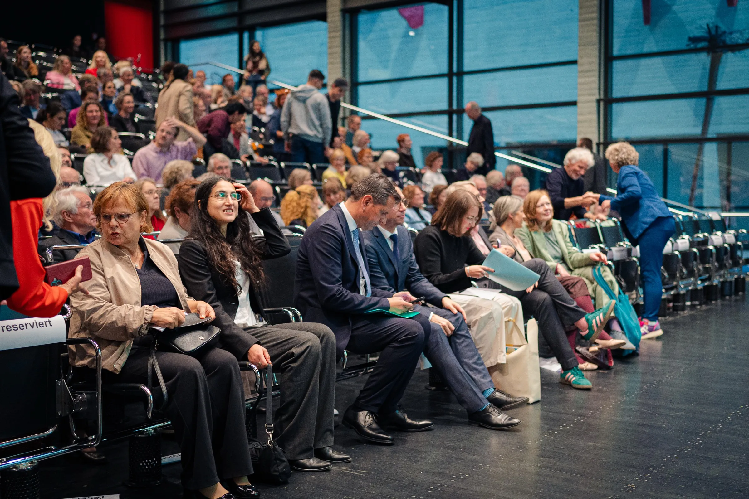 The front row just before the start, guests in suits and blazers, the rising stands in front of large windows behind them