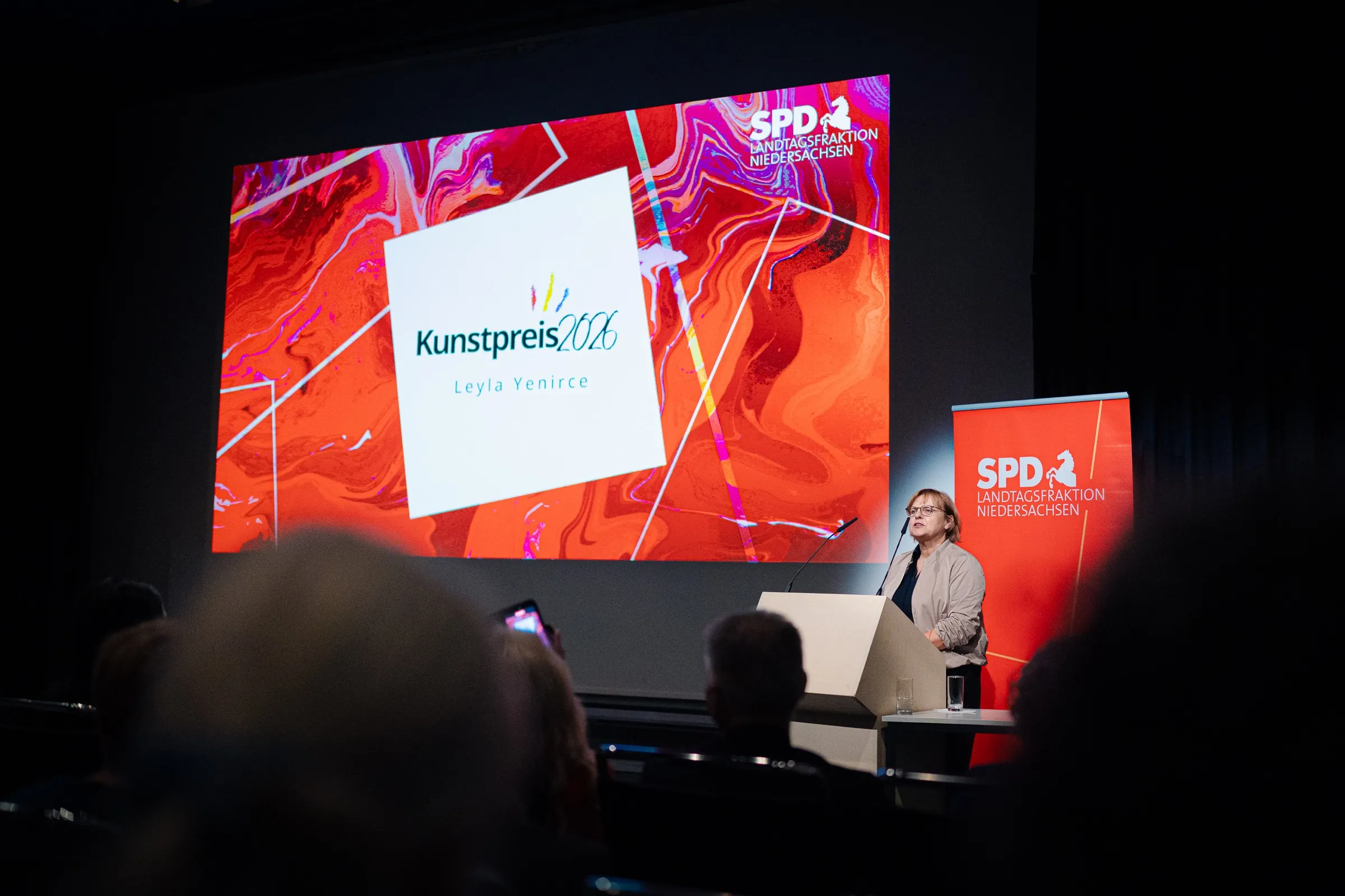 A speaker at the lectern in front of the red and violet wall reading Kunstpreis 2026 and the name Leyla Yenirce