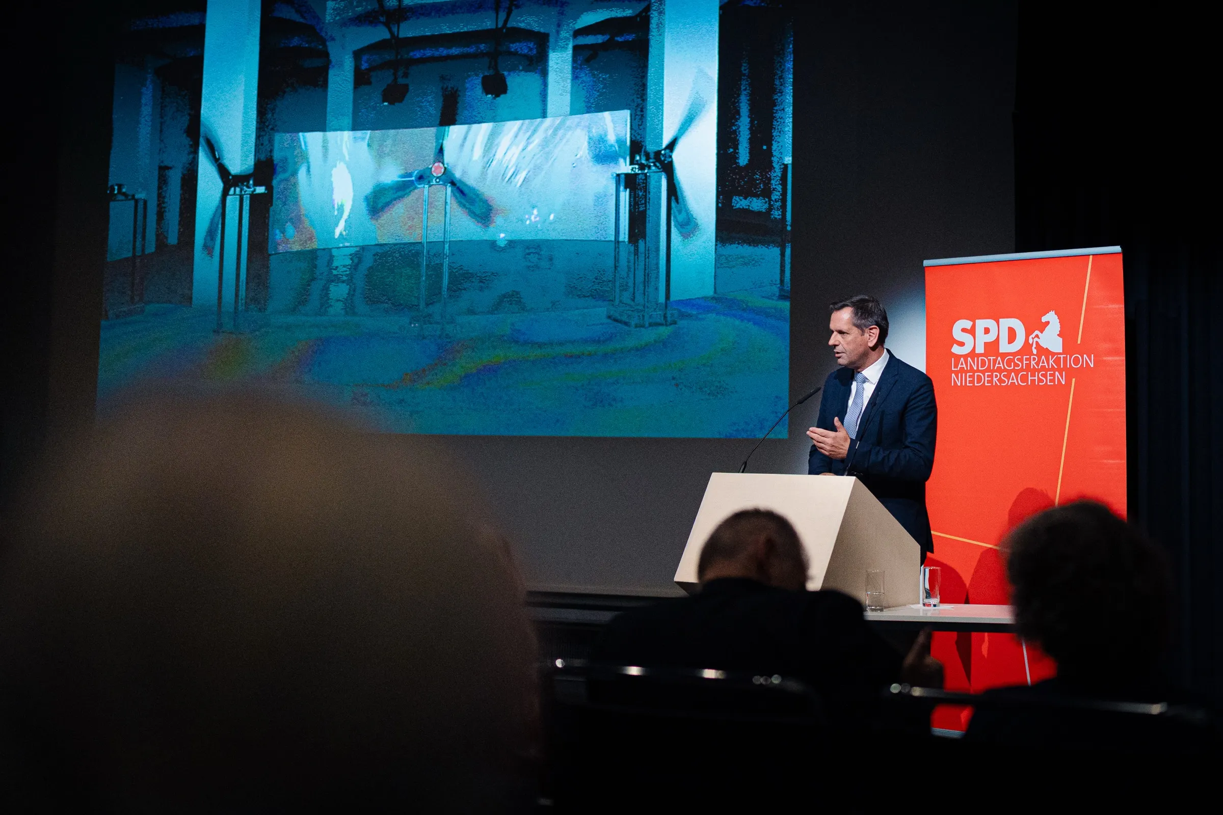 Minister-President Olaf Lies speaking at the lectern, behind him on the screen a video work with large propellers in blue light