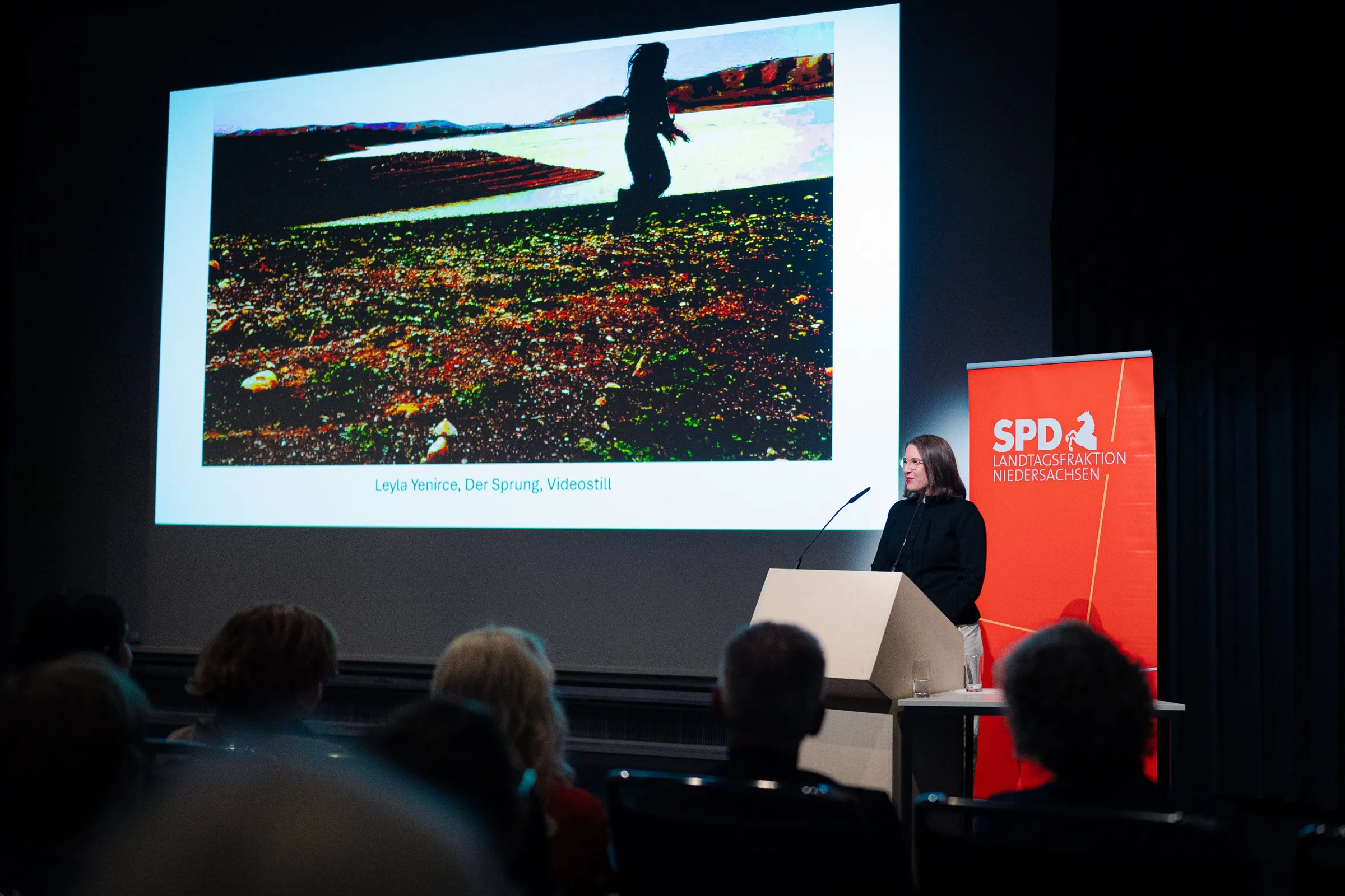 Dr. Anna Heinze at the lectern, on the screen the video still “Der Sprung” captioned Leyla Yenirce, Der Sprung, video still