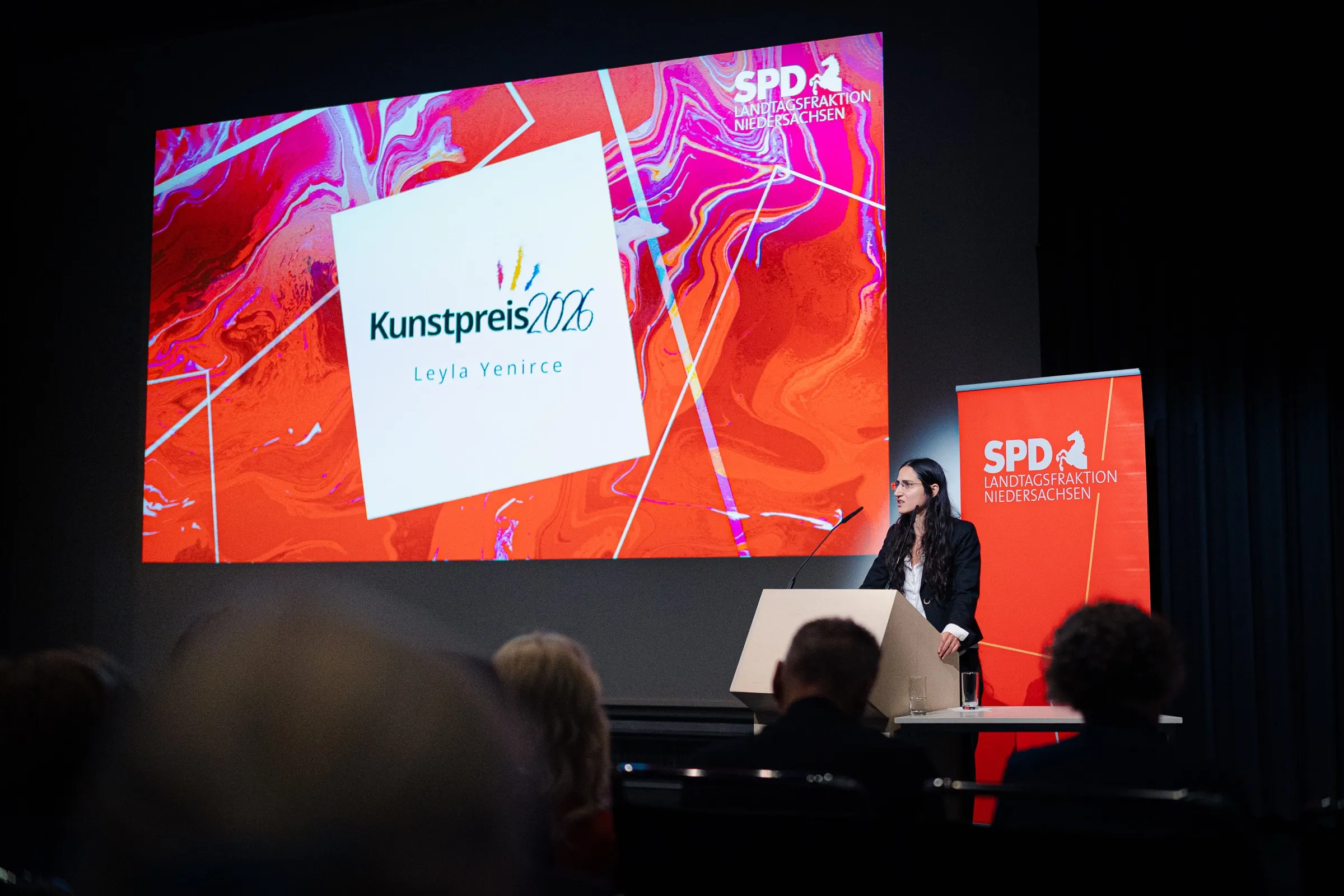 Leyla Yenirce speaking at the lectern in front of the art prize wall