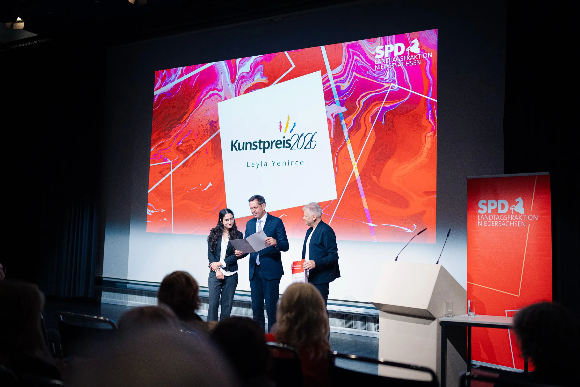 The award on stage in front of the art prize wall: Leyla Yenirce, Olaf Lies and Stefan Politze with the certificate