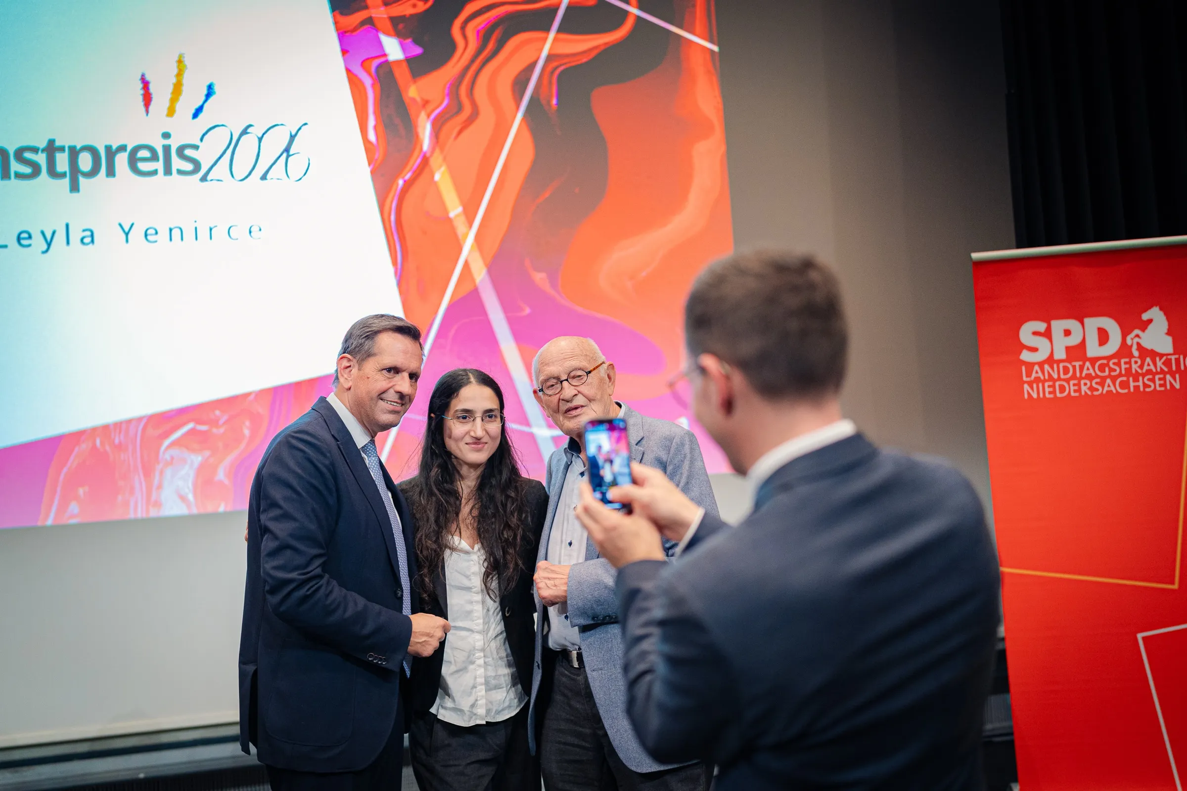 Olaf Lies, Leyla Yenirce and Herbert Schmalstieg posing in front of the art prize wall, in the foreground a man holds up a smartphone