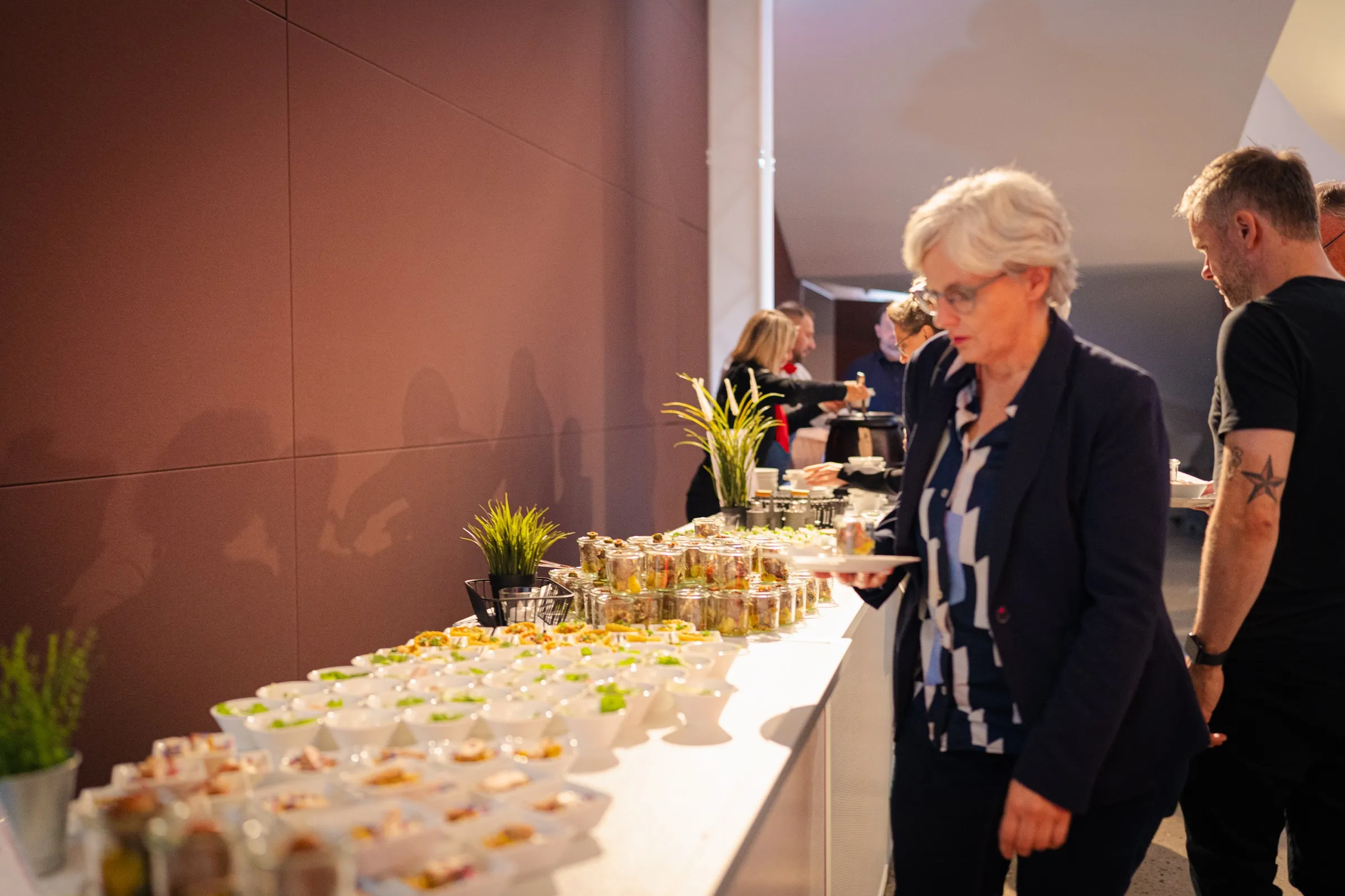 A long counter with small glasses of food and green plants, a woman helping herself