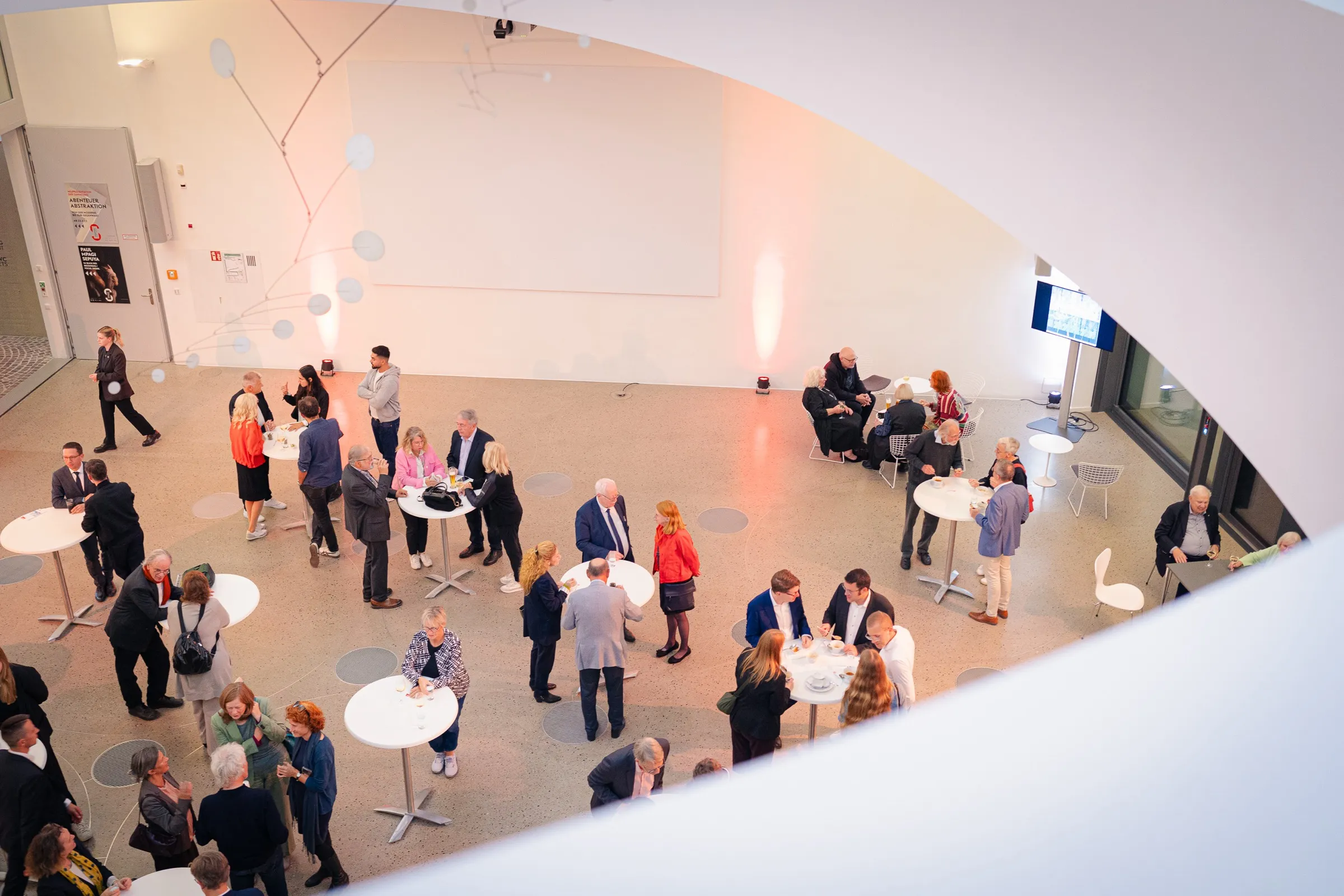 The foyer from above through the curved stairwell opening: standing tables, guests in small groups, a mobile