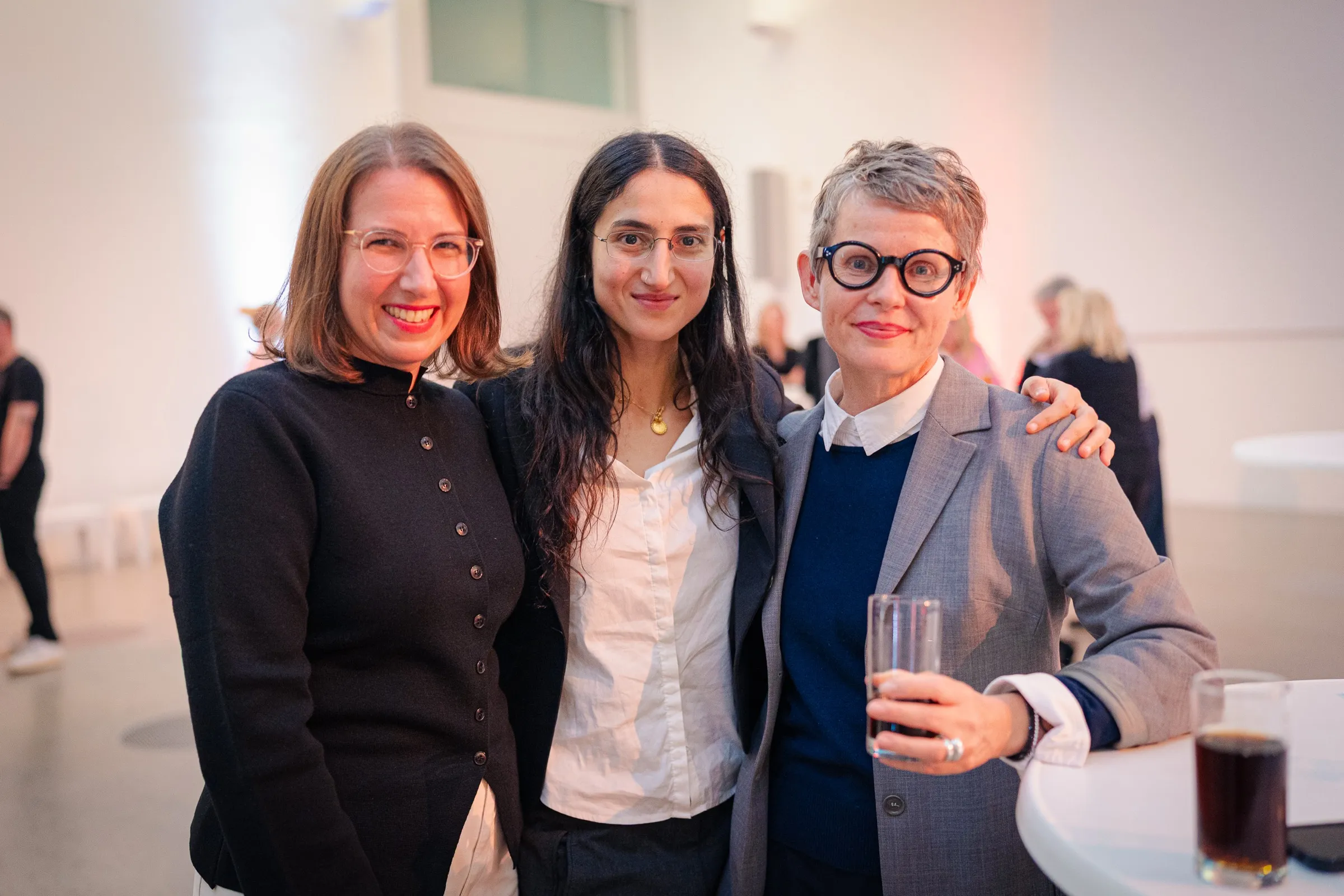 Dr. Anna Heinze, Leyla Yenirce and Dr. Nina Koidl-Weidemann standing arm in arm at a standing table, smiling into the camera