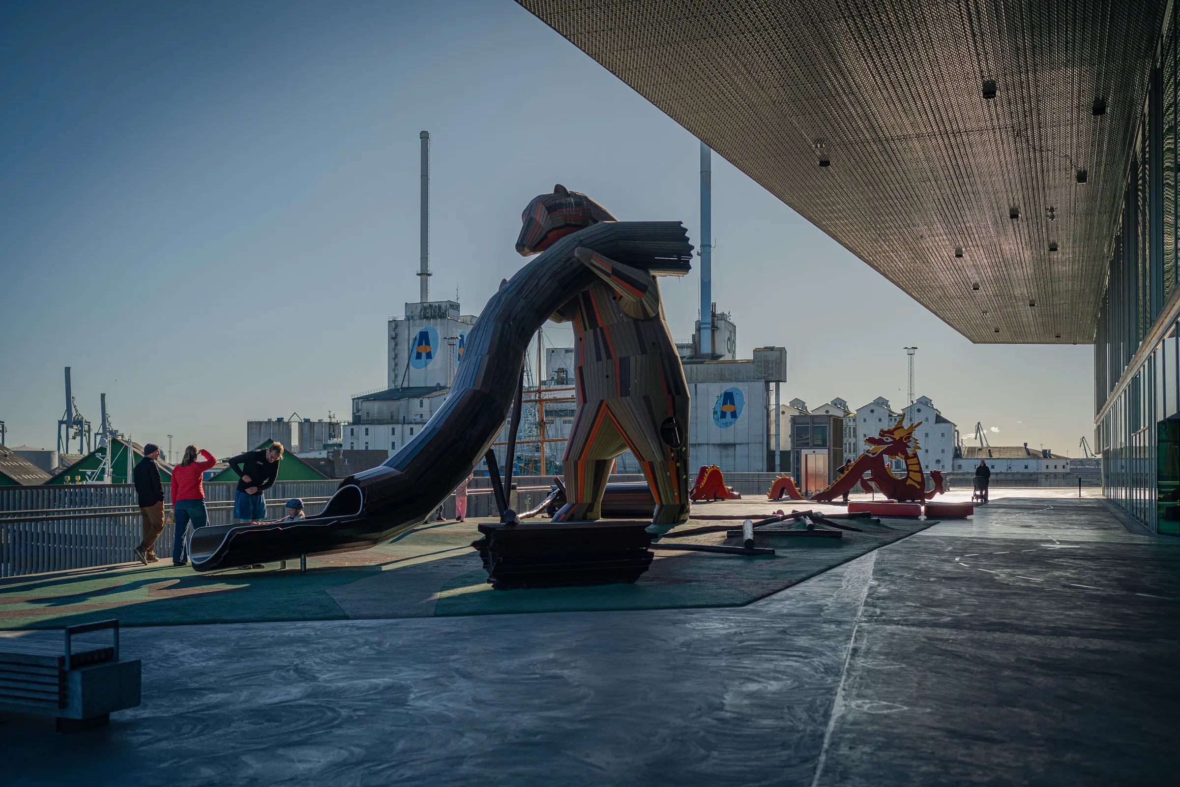 A large animal sculpture on the terrace beneath the roof, harbour, silos and a few people behind