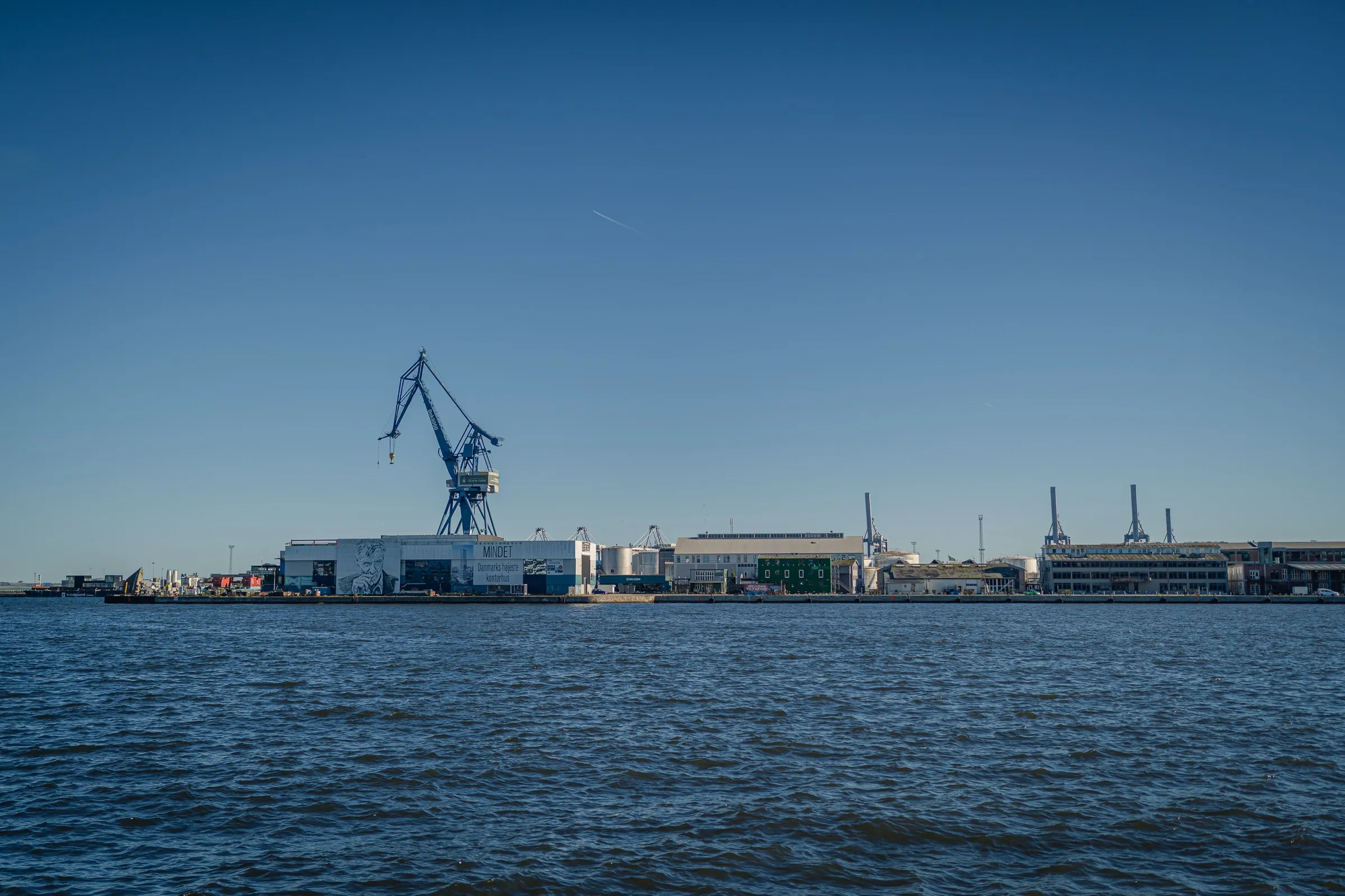 Wide dark blue water, a blue harbour crane and low sheds on the horizon