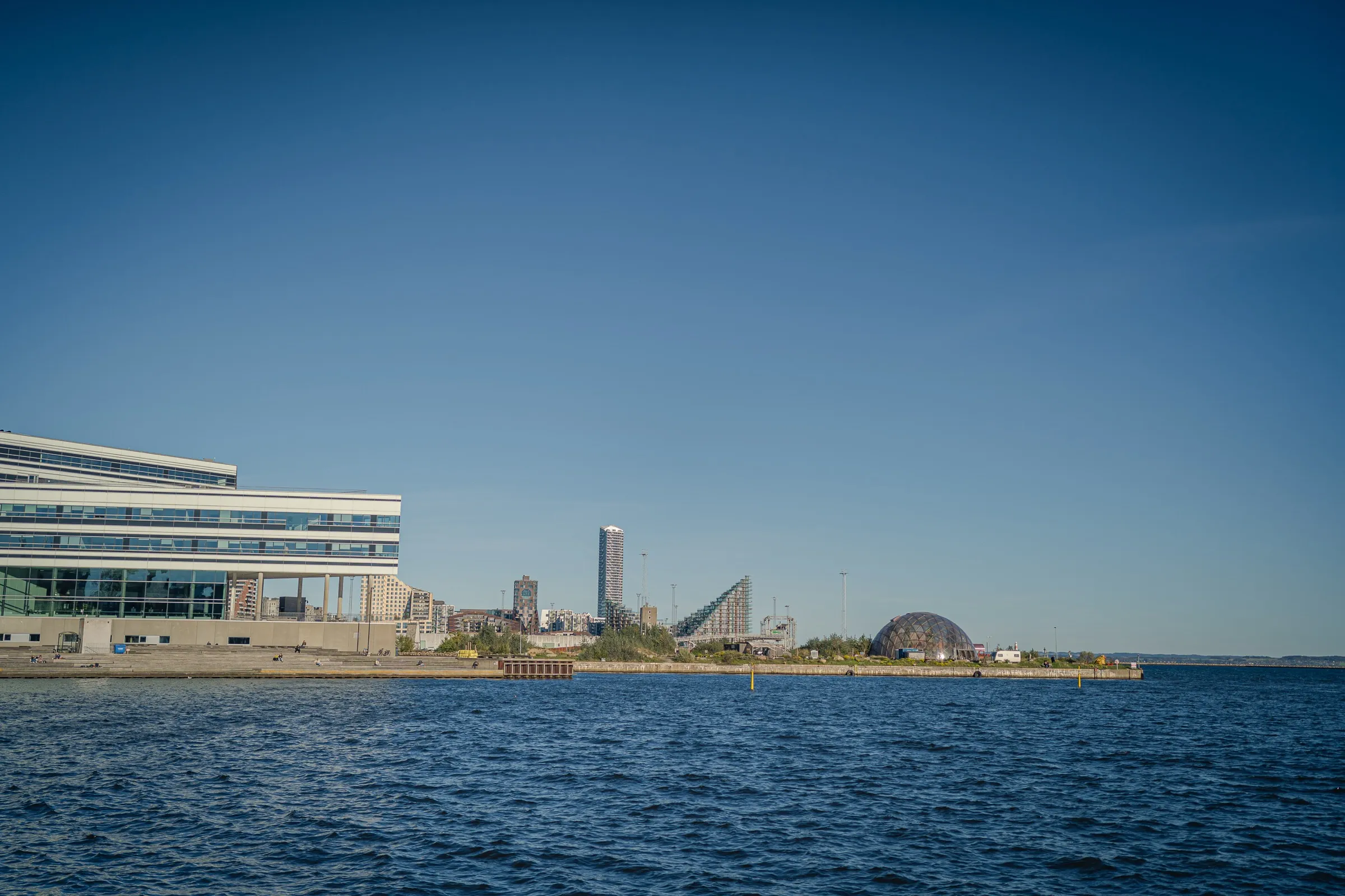 View across the harbour basin to a low building, a residential tower and a dome on the shore